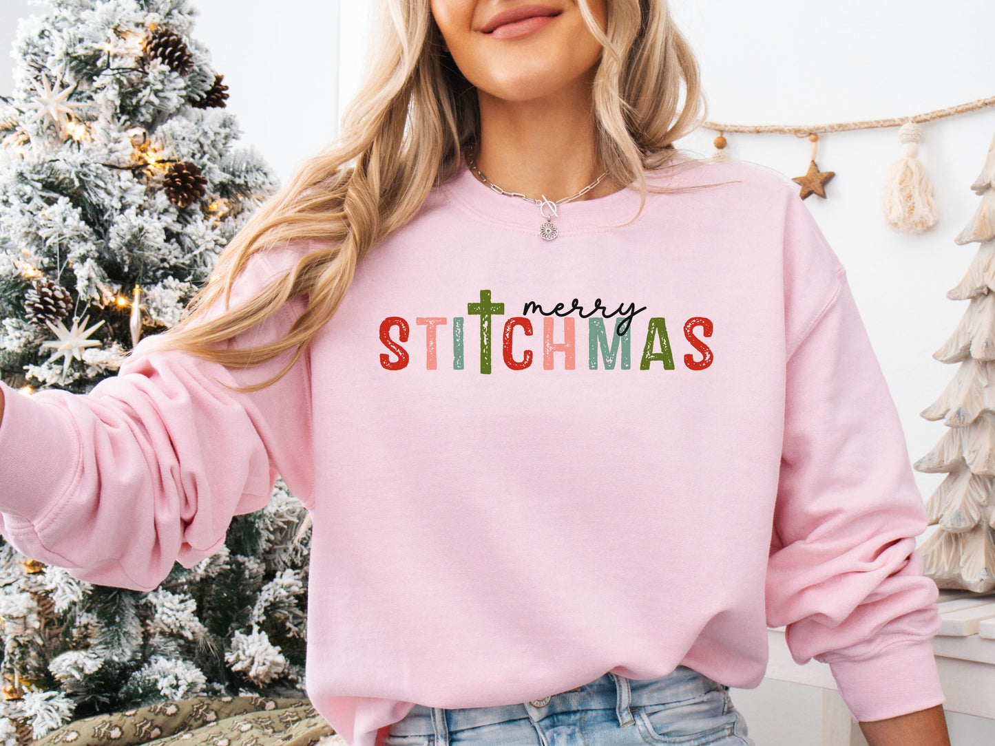 Lt-Pink colored long sleeve shirt with the phrase “Merry Stitchmas” featuring a cross-shaped “T,” styled in a cozy Christmas setting with festive décor and sewing details.
