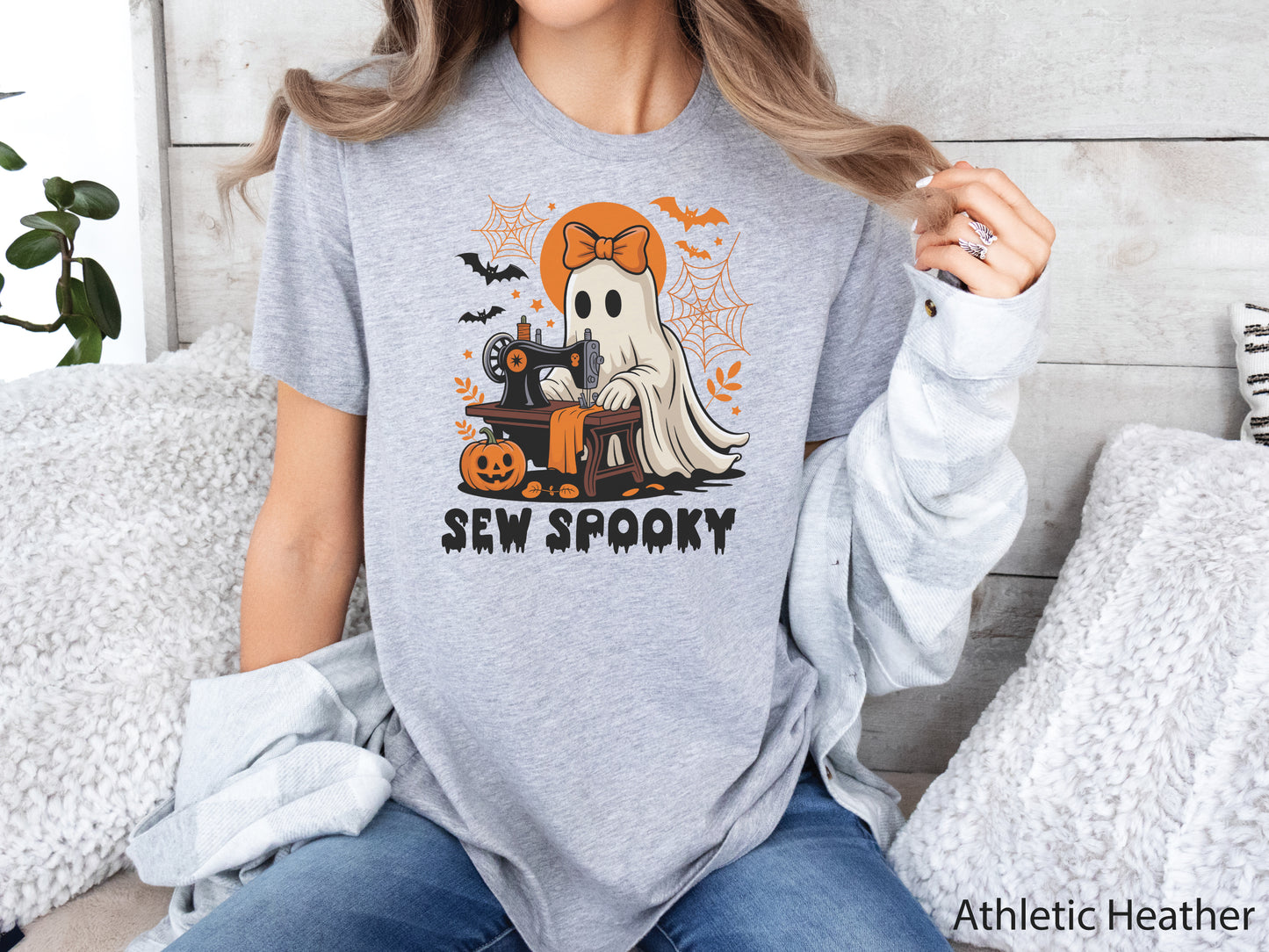Athletic Heather color t-shirt with a Halloween sewing design featuring a ghost at a vintage sewing machine, surrounded by pumpkins, bats, spiderwebs, and stars, with the phrase “Sew Spooky” in bold black lettering.