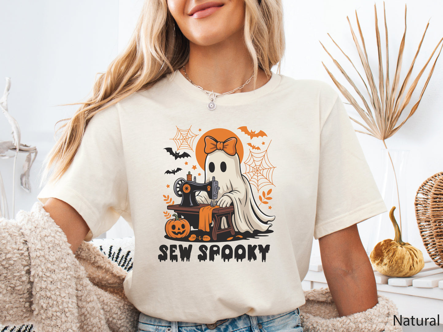 Natural color t-shirt with a Halloween sewing design featuring a ghost at a vintage sewing machine, surrounded by pumpkins, bats, spiderwebs, and stars, with the phrase “Sew Spooky” in bold black lettering.
