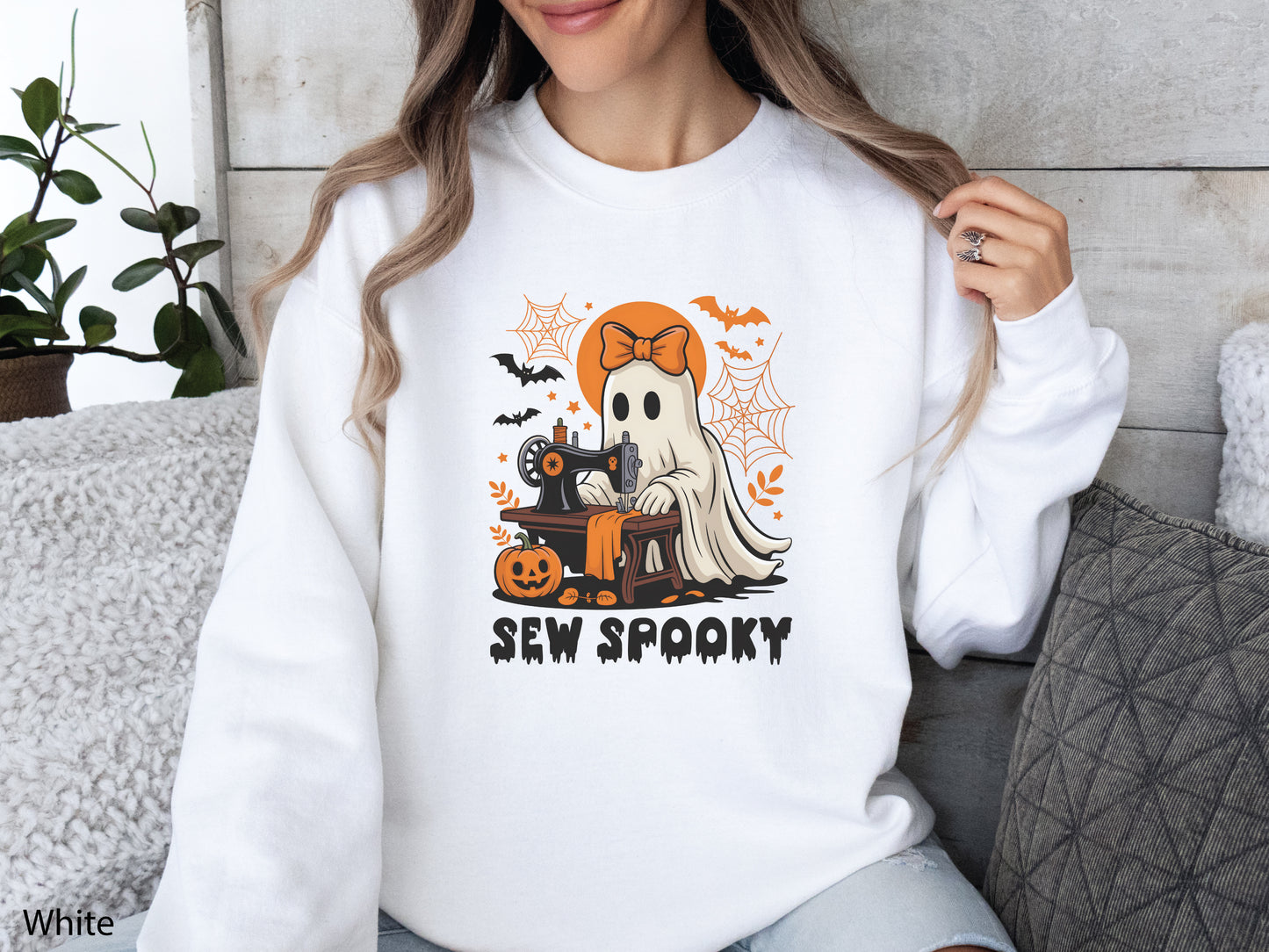 Cozy White sweatshirt with a Halloween sewing design featuring a ghost at a vintage sewing machine, surrounded by pumpkins, bats, spiderwebs, and stars, with the phrase “Sew Spooky” in bold black lettering.