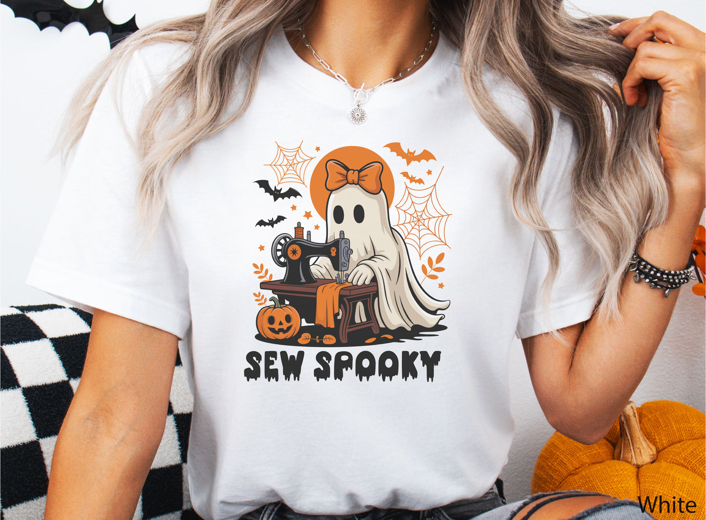 White color t-shirt with a Halloween sewing design featuring a ghost at a vintage sewing machine, surrounded by pumpkins, bats, spiderwebs, and stars, with the phrase “Sew Spooky” in bold black lettering.