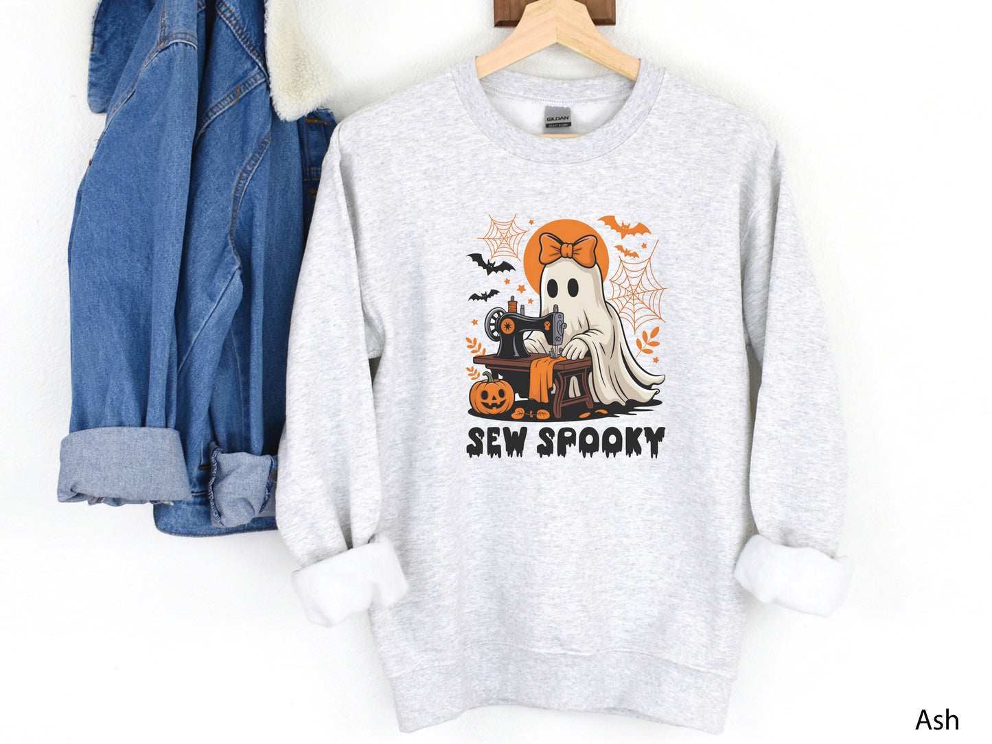 Cozy light grey color sweatshirt with a Halloween sewing design featuring a ghost at a vintage sewing machine, surrounded by pumpkins, bats, spiderwebs, and stars, with the phrase “Sew Spooky” in bold black lettering.