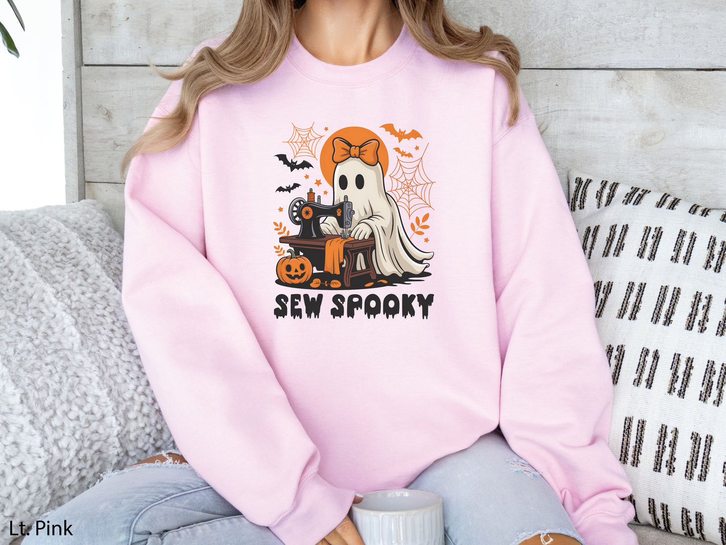 Cozy beige light pink color sweatshirt with a Halloween sewing design featuring a ghost at a vintage sewing machine, surrounded by pumpkins, bats, spiderwebs, and stars, with the phrase “Sew Spooky” in bold black lettering.