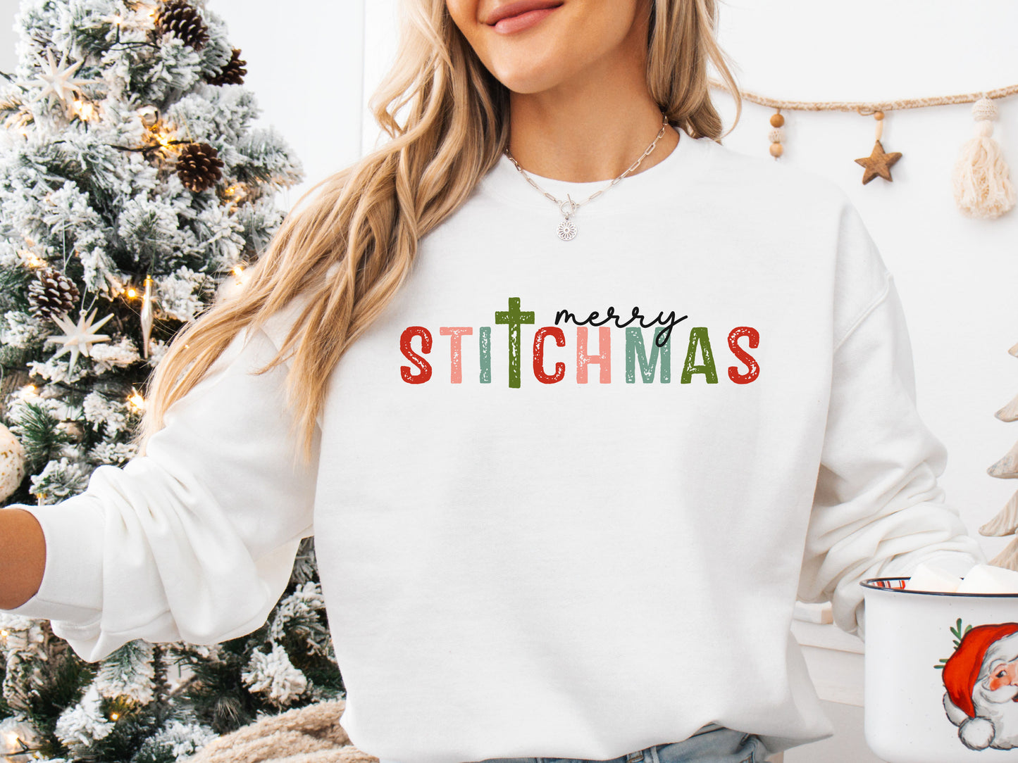 White colored long sleeve shirt with the phrase “Merry Stitchmas” featuring a cross-shaped “T,” styled in a cozy Christmas setting with festive décor and sewing details.
