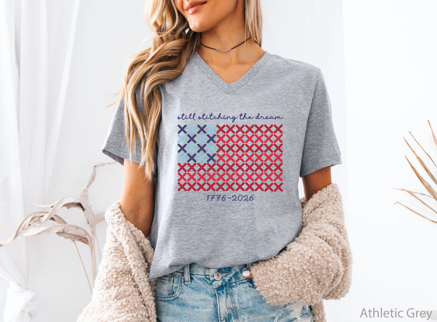 Patriotic v-neck t-shirt with cross-stitch American flag design and the phrase Still Stitching the Dream 1776–2026, ideal for quilters and needlework enthusiasts.