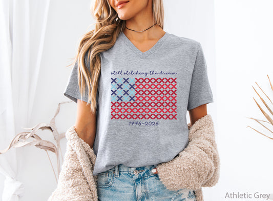 Patriotic v-neck t-shirt with cross-stitch American flag design and the phrase Still Stitching the Dream 1776–2026, ideal for quilters and needlework enthusiasts.