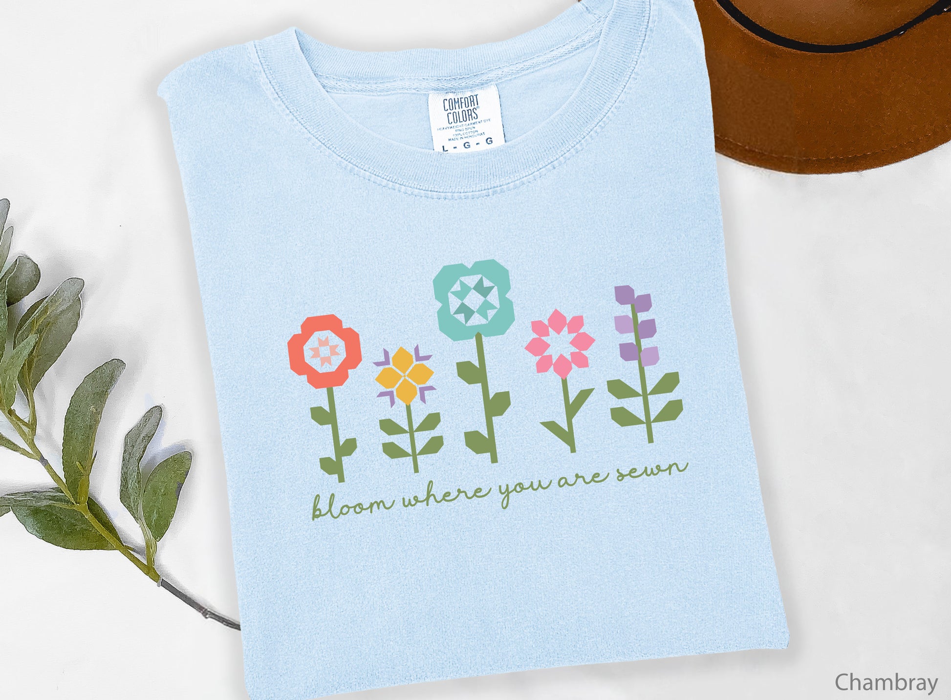 Light blue (Chambray) t-shirt featuring colorful quilt block flowers with green stems and the phrase "Bloom Where You Are Sewn," perfect for quilters and sewing enthusiasts.