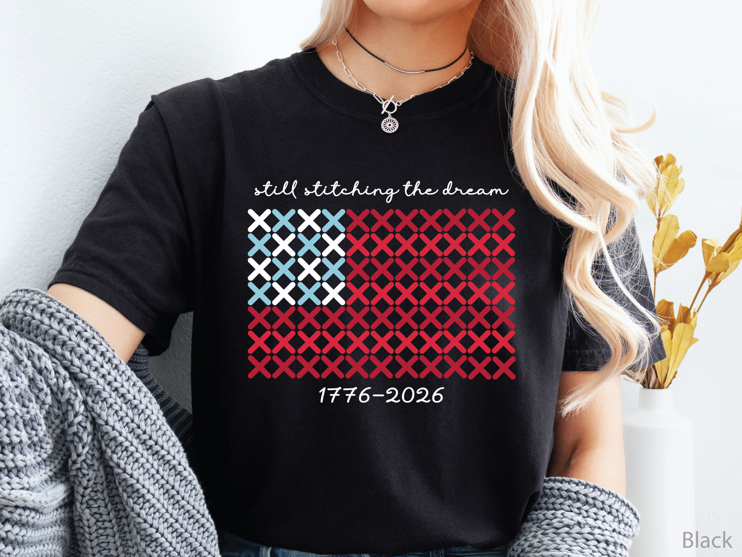 Patriotic crew t-shirt with cross-stitch American flag design and the phrase Still Stitching the Dream 1776–2026, ideal for quilters and needlework enthusiasts.