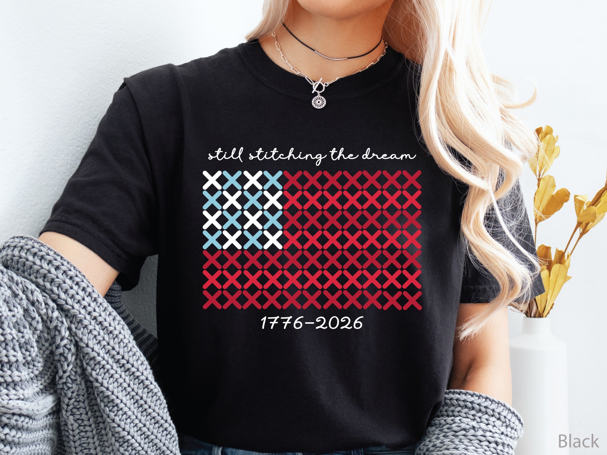 Patriotic crew t-shirt with cross-stitch American flag design and the phrase Still Stitching the Dream 1776–2026, ideal for quilters and needlework enthusiasts.