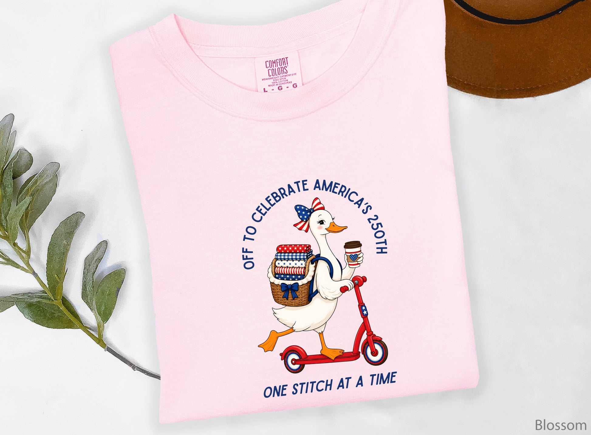 Patriotic quilting t-shirt design featuring a goose riding a red scooter with a basket of red, white, and blue quilt fabrics, paired with the phrases “Off To Celebrate America’s 250th” and “One Quilt At A Time.”