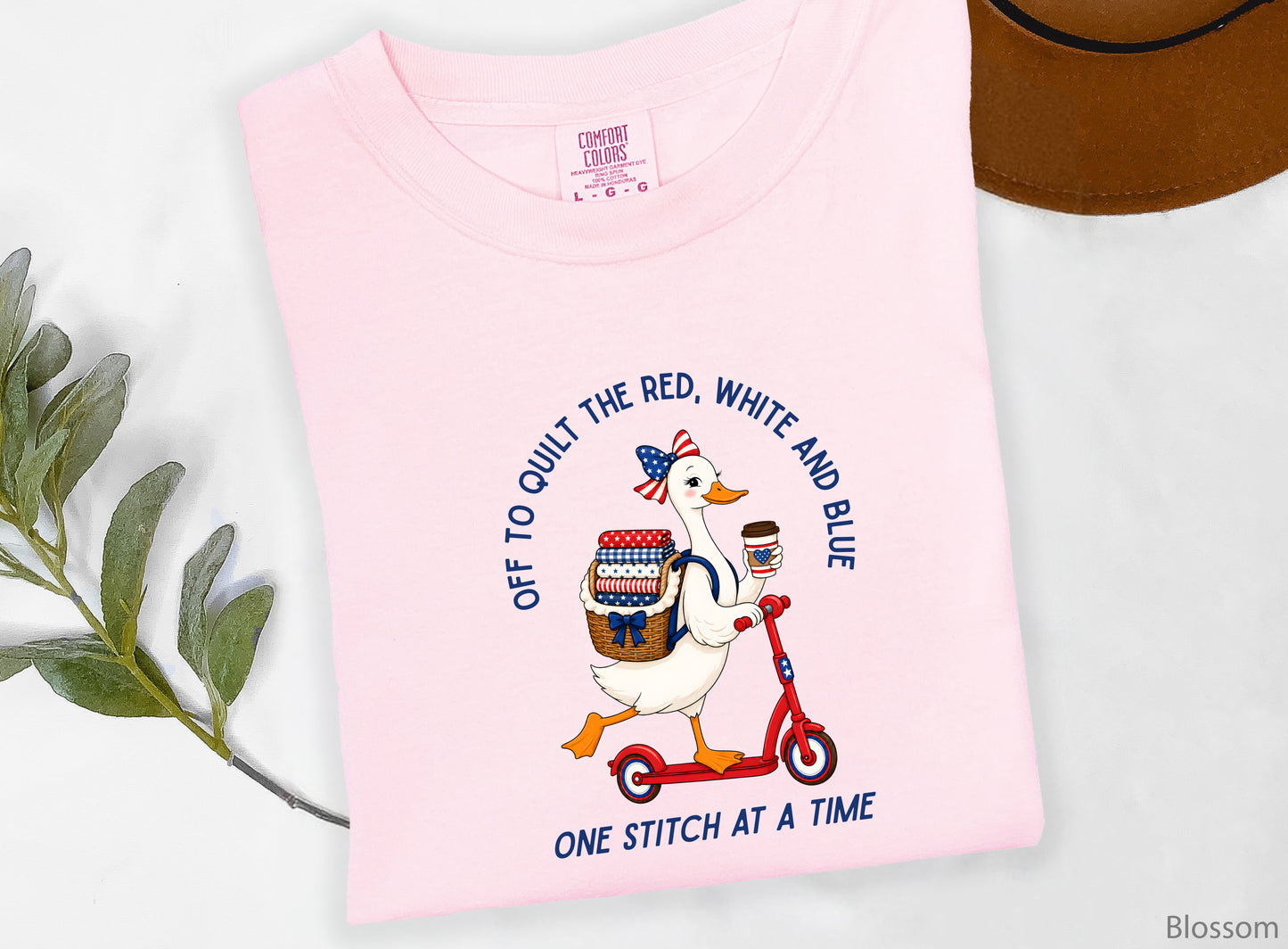 Patriotic quilting t-shirt design featuring a goose riding a red scooter with a basket of red, white, and blue quilt fabrics, paired with the phrases “Off To Stitch The Red, White and Blue” and “One Quilt At A Time.”