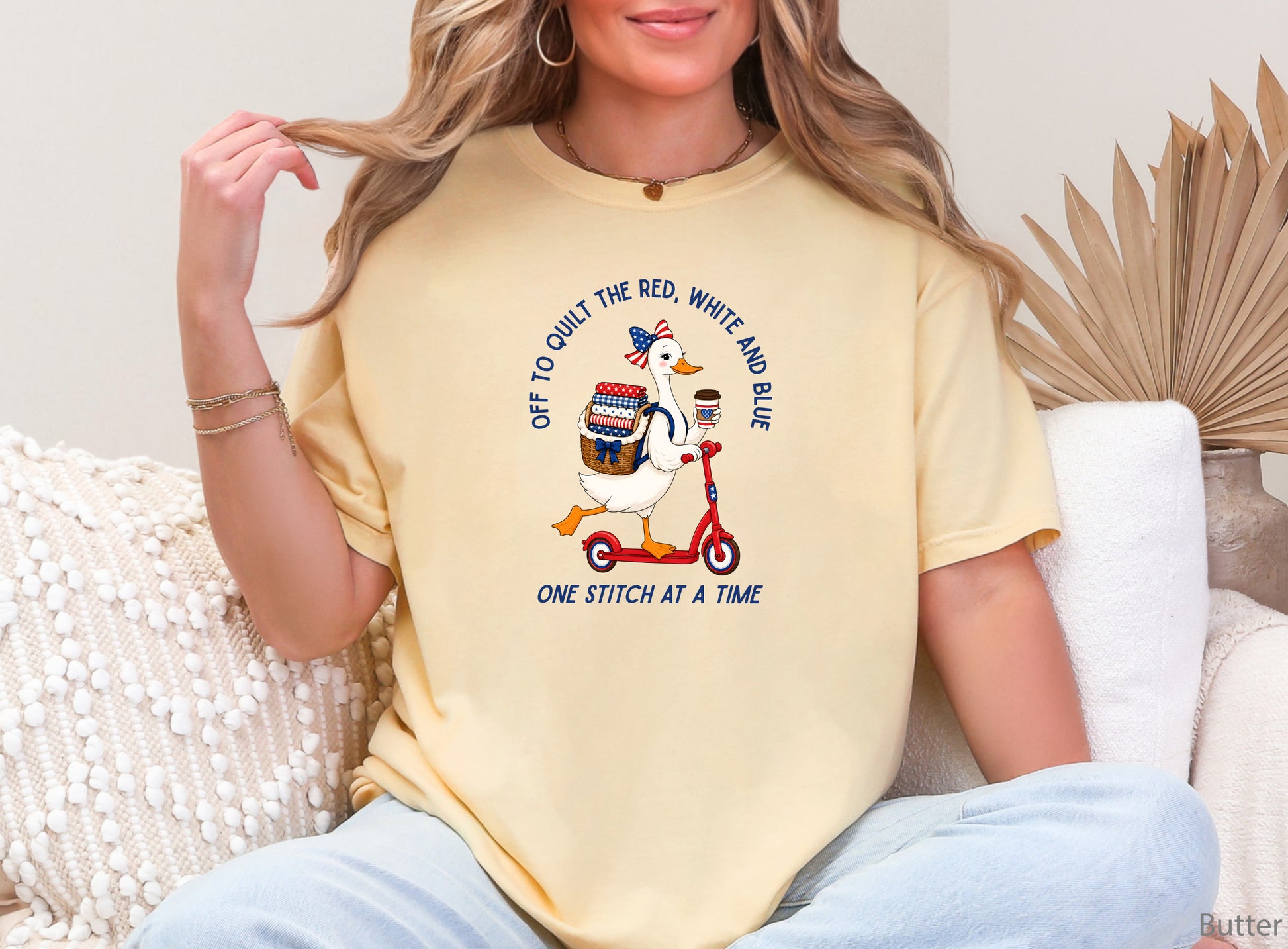 Patriotic quilting t-shirt design featuring a goose riding a red scooter with a basket of red, white, and blue quilt fabrics, paired with the phrases “Off To Stitch The Red, White and Blue” and “One Quilt At A Time.”