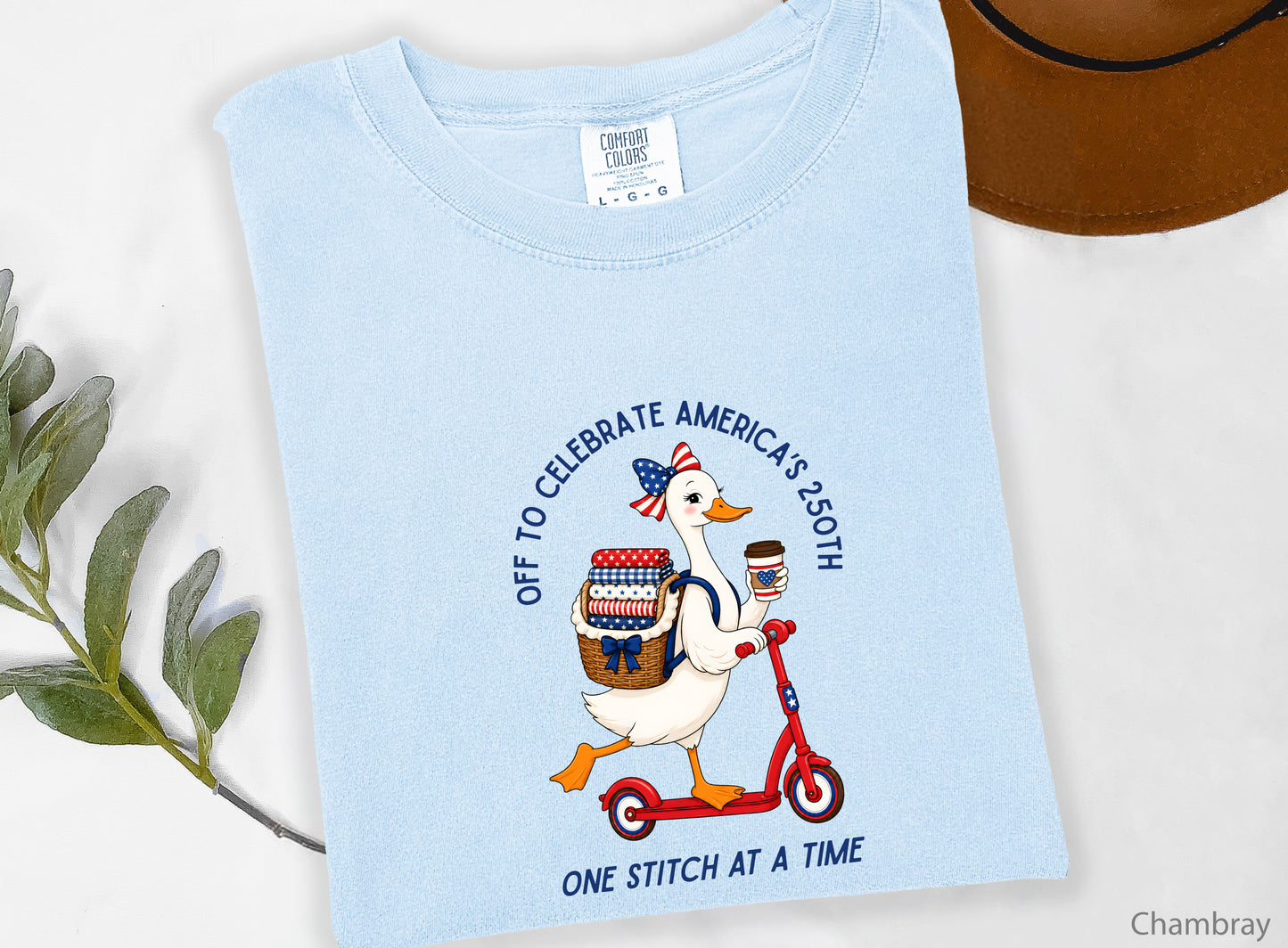 Patriotic quilting t-shirt design featuring a goose riding a red scooter with a basket of red, white, and blue quilt fabrics, paired with the phrases “Off To Celebrate America’s 250th” and “One Quilt At A Time.”
