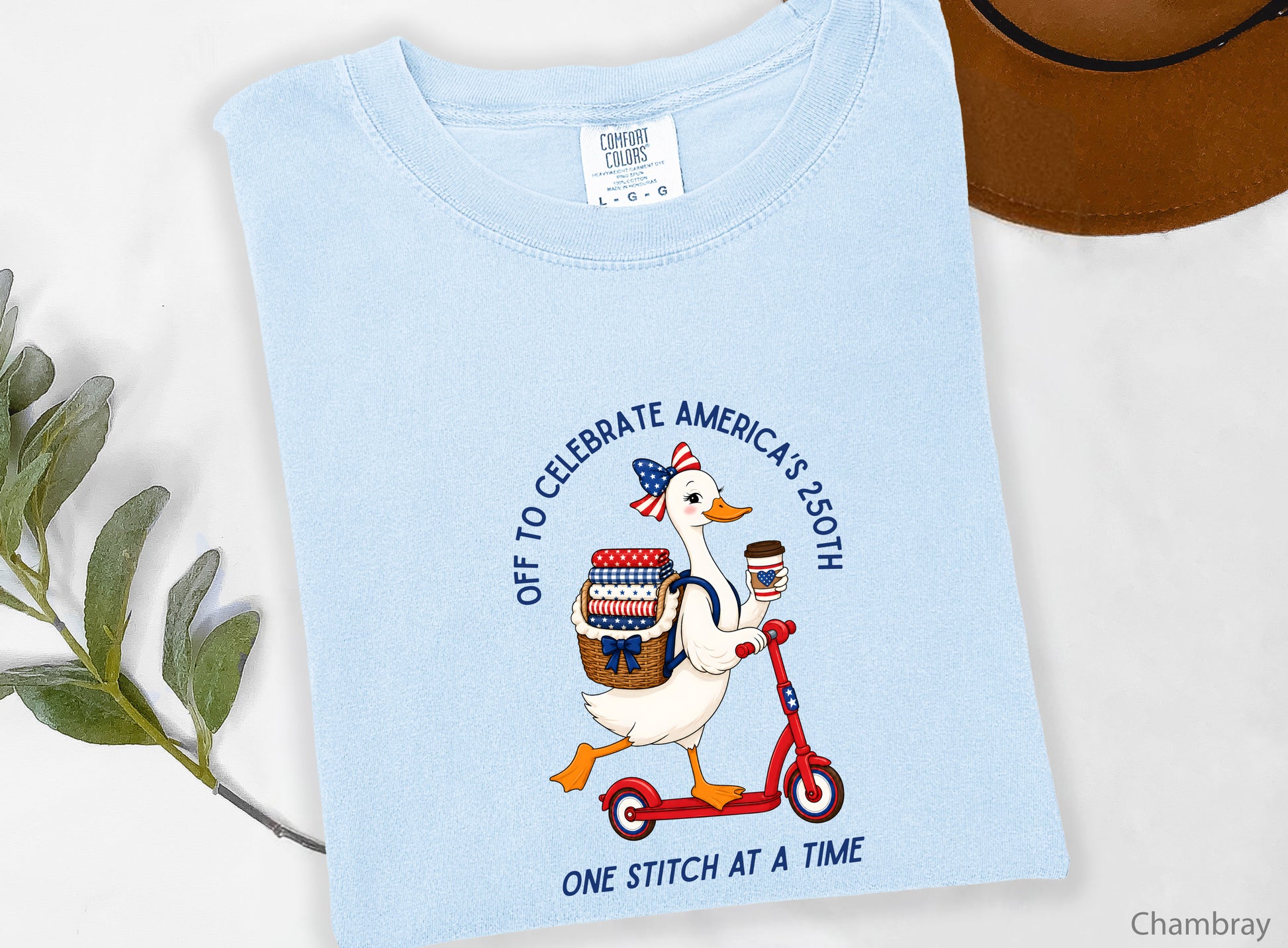 Patriotic quilting t-shirt design featuring a goose riding a red scooter with a basket of red, white, and blue quilt fabrics, paired with the phrases “Off To Celebrate America’s 250th” and “One Quilt At A Time.”