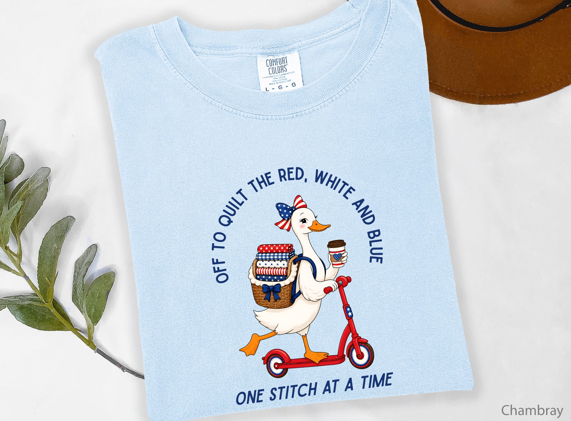 Patriotic quilting t-shirt design featuring a goose riding a red scooter with a basket of red, white, and blue quilt fabrics, paired with the phrases “Off To Stitch The Red, White and Blue” and “One Quilt At A Time.”