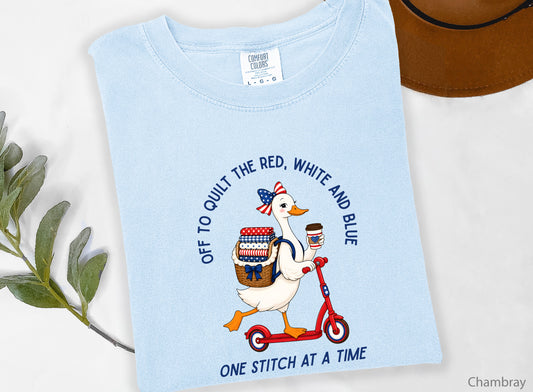 Patriotic quilting t-shirt design featuring a goose riding a red scooter with a basket of red, white, and blue quilt fabrics, paired with the phrases “Off To Stitch The Red, White and Blue” and “One Quilt At A Time.”