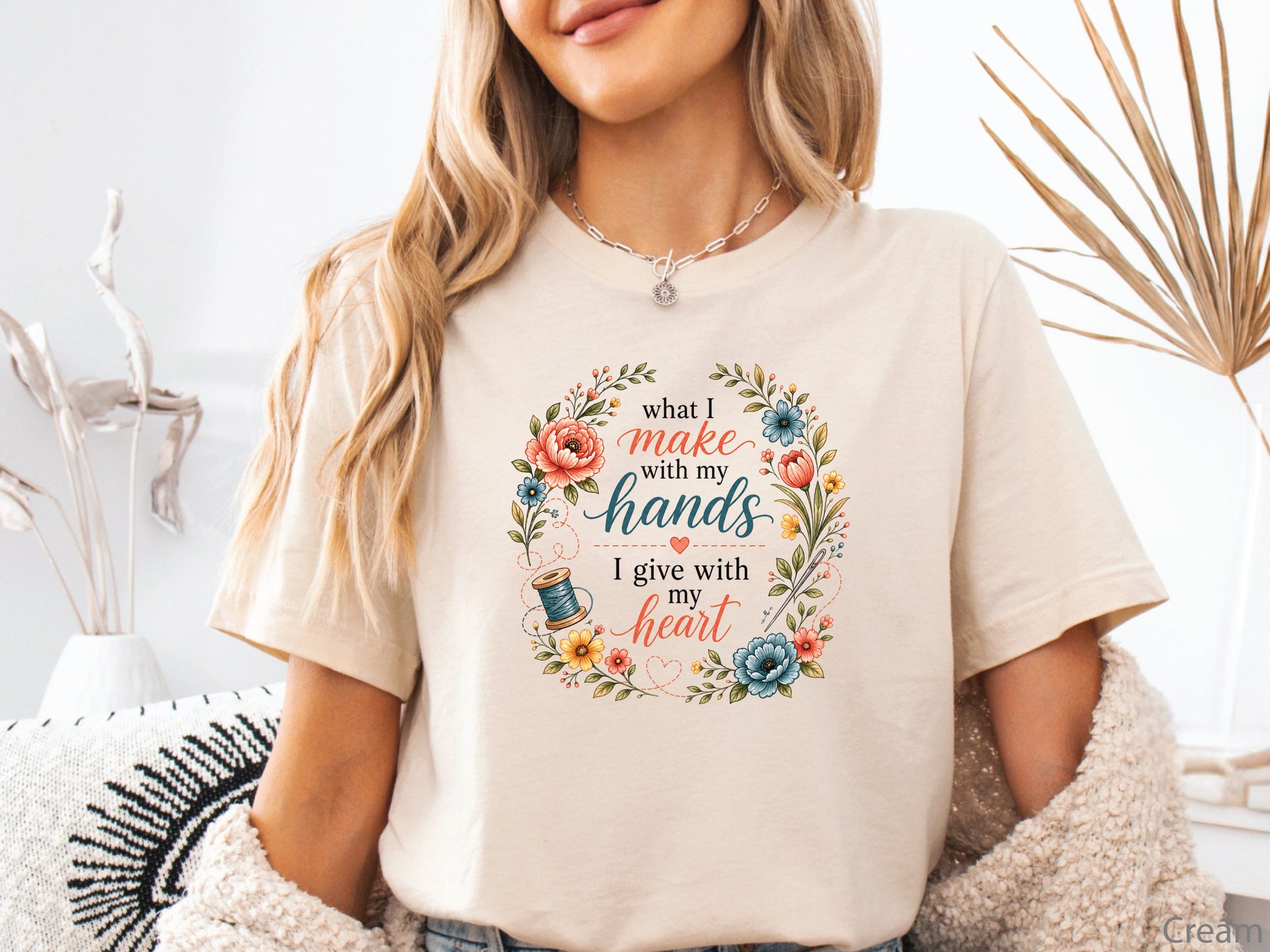 Inspirational sewing t-shirt featuring the phrase "What I Make With My Hands, I Give With My Heart" surrounded by a floral wreath with a spool of thread and stitched accents, perfect for sewists and quilters.