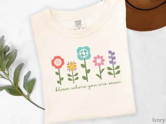 Ivory t-shirt featuring colorful quilt block flowers with green stems and the phrase "Bloom Where You Are Sewn," perfect for quilters and sewing enthusiasts.