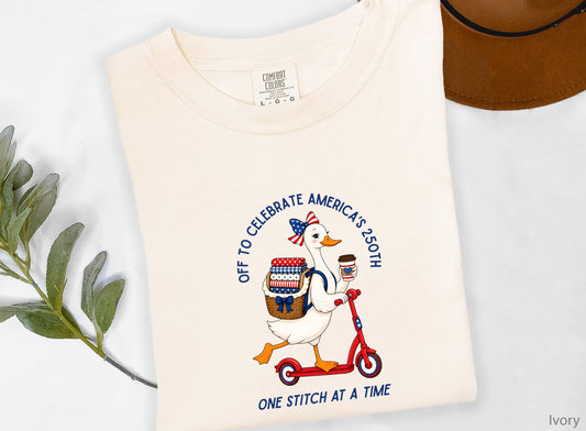 Patriotic quilting t-shirt design featuring a goose riding a red scooter with a basket of red, white, and blue quilt fabrics, paired with the phrases “Off To Celebrate America’s 250th” and “One Quilt At A Time.”