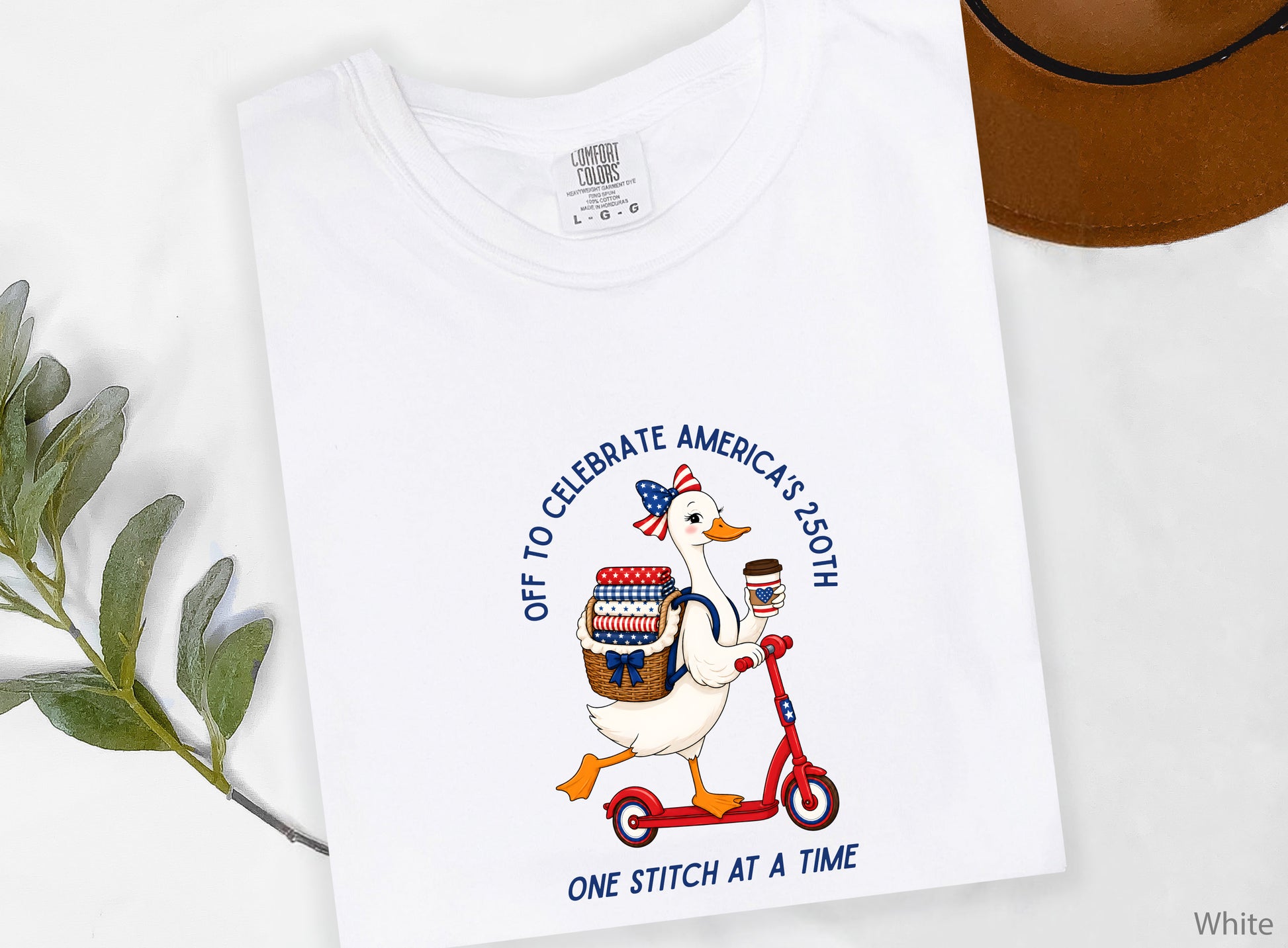 Patriotic quilting t-shirt design featuring a goose riding a red scooter with a basket of red, white, and blue quilt fabrics, paired with the phrases “Off To Celebrate America’s 250th” and “One Quilt At A Time.”