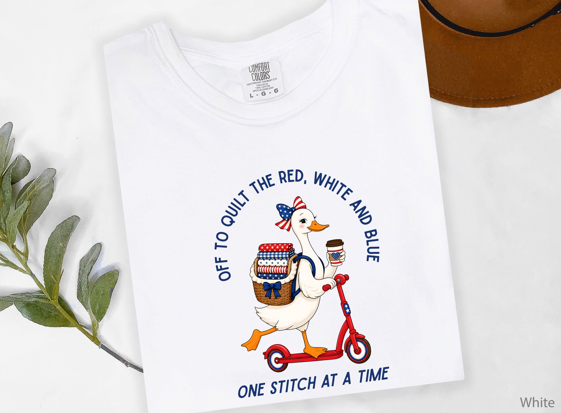 Patriotic quilting t-shirt design featuring a goose riding a red scooter with a basket of red, white, and blue quilt fabrics, paired with the phrases “Off To Stitch The Red, White and Blue” and “One Quilt At A Time.”