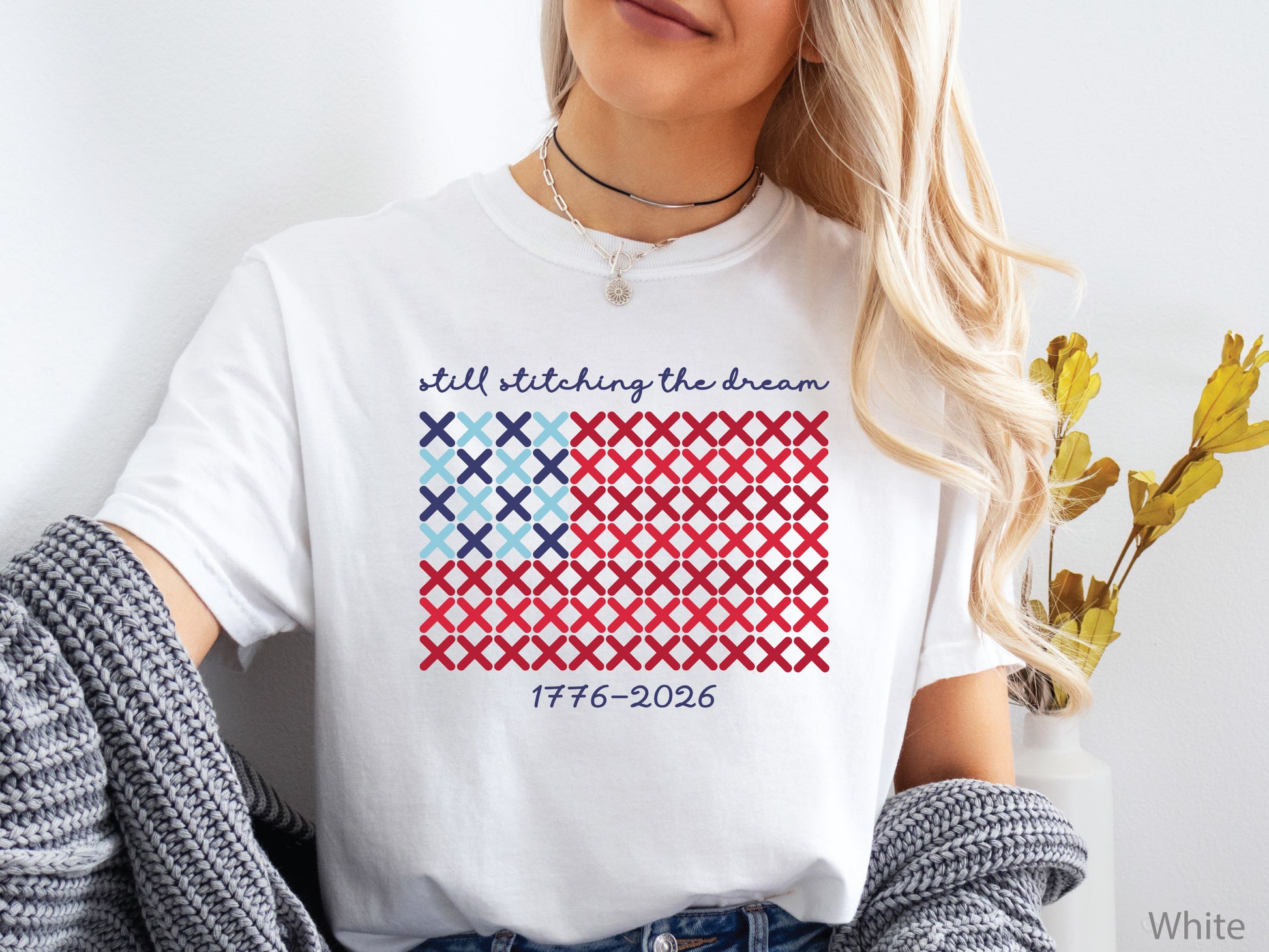 Patriotic crew t-shirt with cross-stitch American flag design and the phrase Still Stitching the Dream 1776–2026, ideal for quilters and needlework enthusiasts.