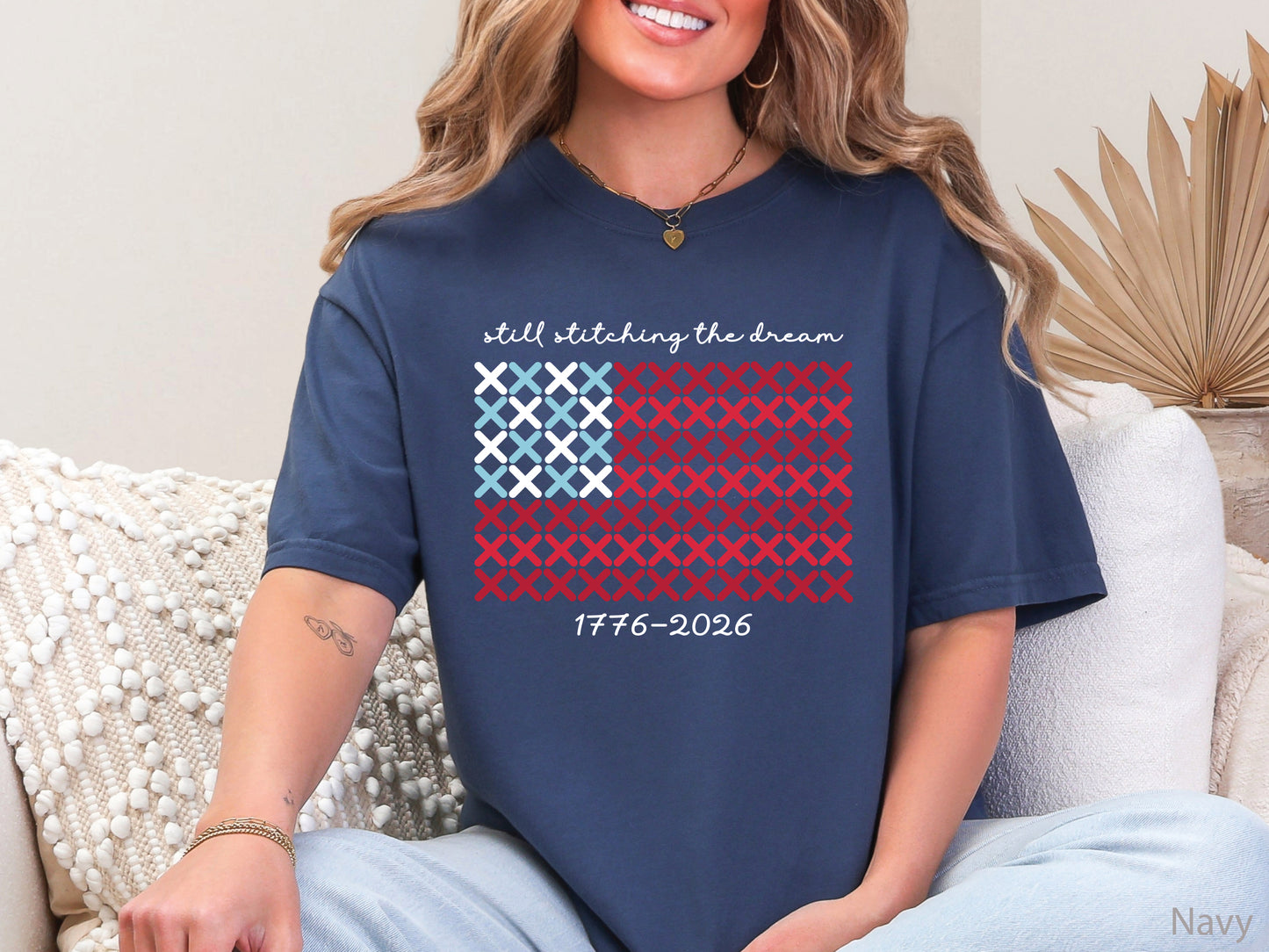 Patriotic crew t-shirt with cross-stitch American flag design and the phrase Still Stitching the Dream 1776–2026, ideal for quilters and needlework enthusiasts.