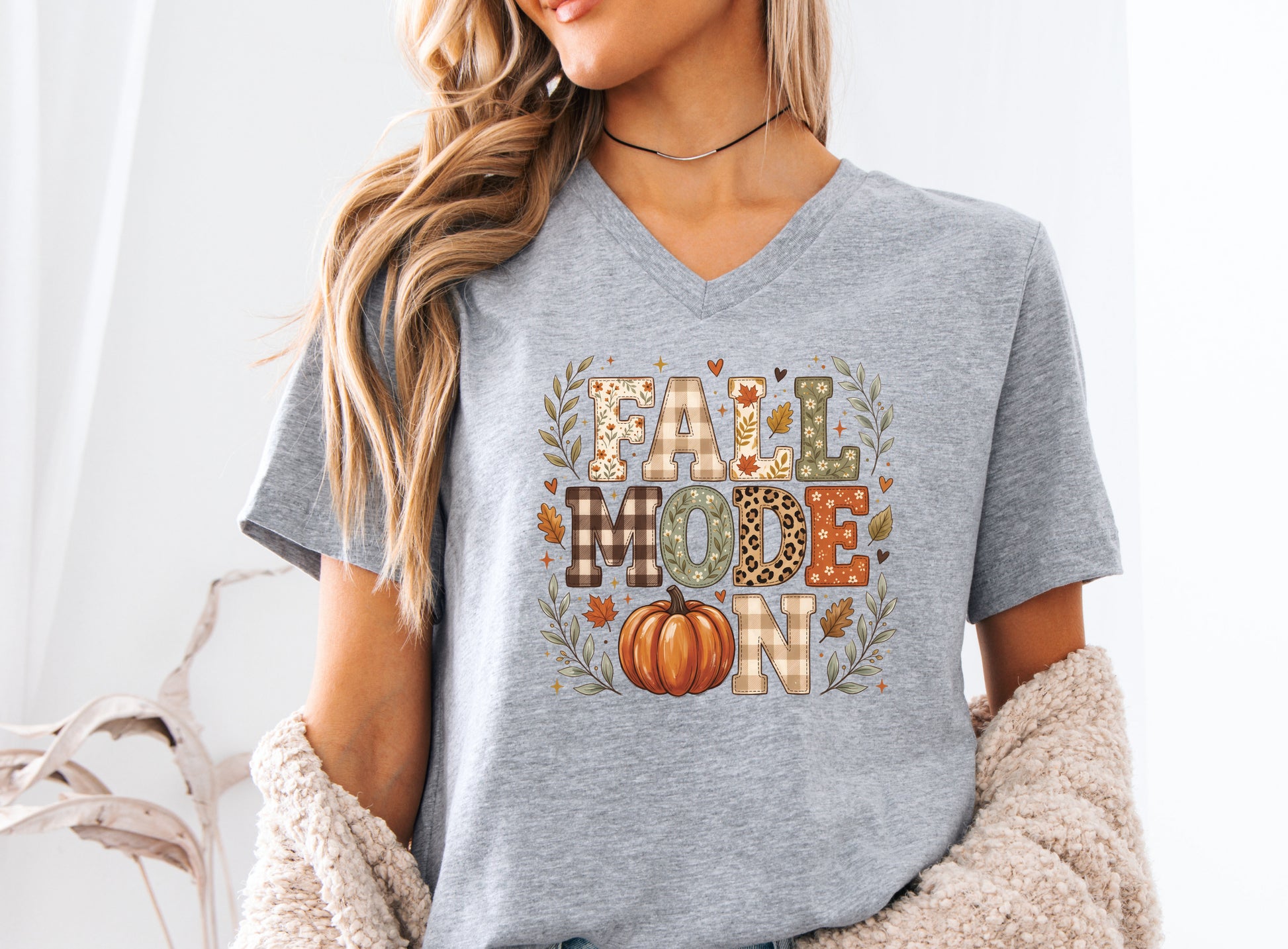 Fall Mode On v-neck t-shirt design featuring patchwork-style lettering with patchwork plaid, floral, leopard print, and fall leaf patterns, plus an orange pumpkin and autumn foliage.