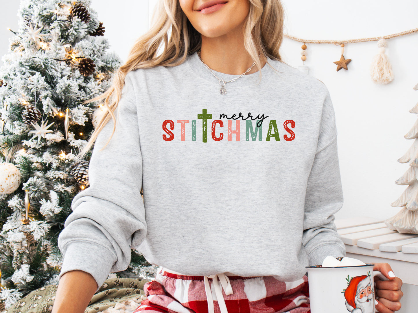 Ash (grey) colored long sleeve shirt with the phrase “Merry Stitchmas” featuring a cross-shaped “T,” styled in a cozy Christmas setting with festive décor and sewing details.