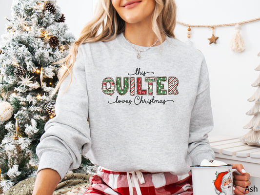 Ash (light grey) sweatshirt with the phrase “This Quilter Loves Christmas” in red and green patchwork-patterned letters, styled in a cozy holiday setting with festive décor.