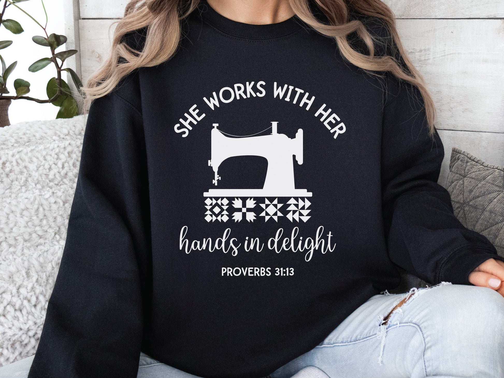 A crew style sweatshirt featuring a vintage-style sewing machine, quilt block icons, and the verse “She works with her hands in delight – Proverbs 31:13.”