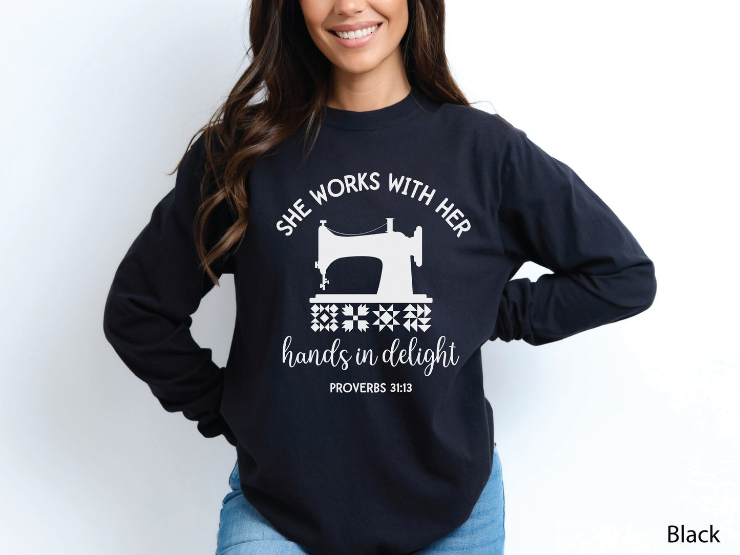 A long-sleeve  t-shirt featuring a vintage-style sewing machine, quilt block icons, and the verse “She works with her hands in delight – Proverbs 31:13.”