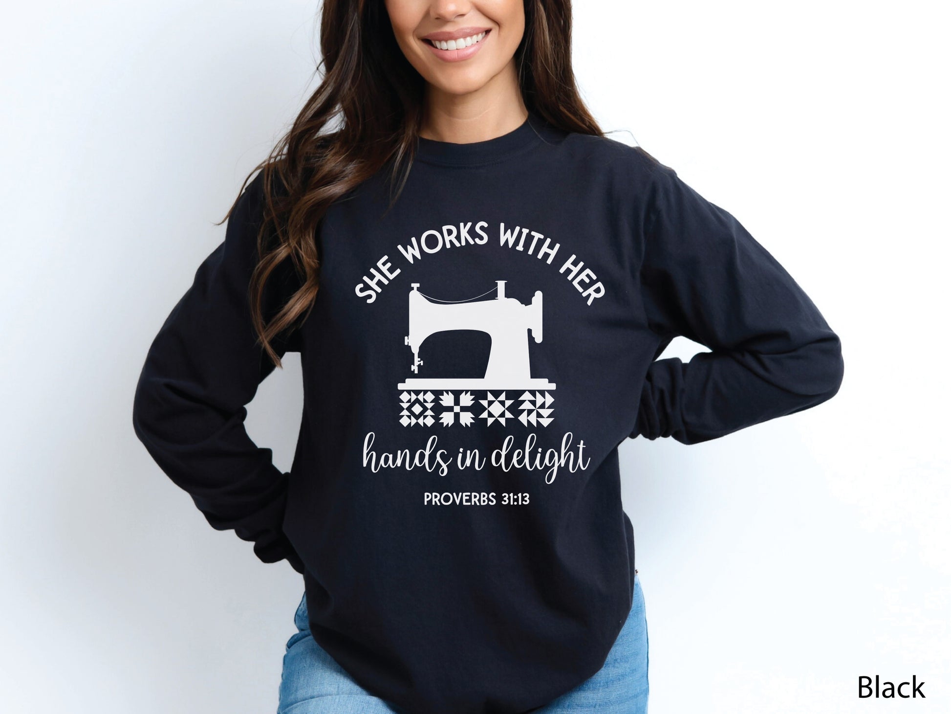 A long-sleeve  t-shirt featuring a vintage-style sewing machine, quilt block icons, and the verse “She works with her hands in delight – Proverbs 31:13.”