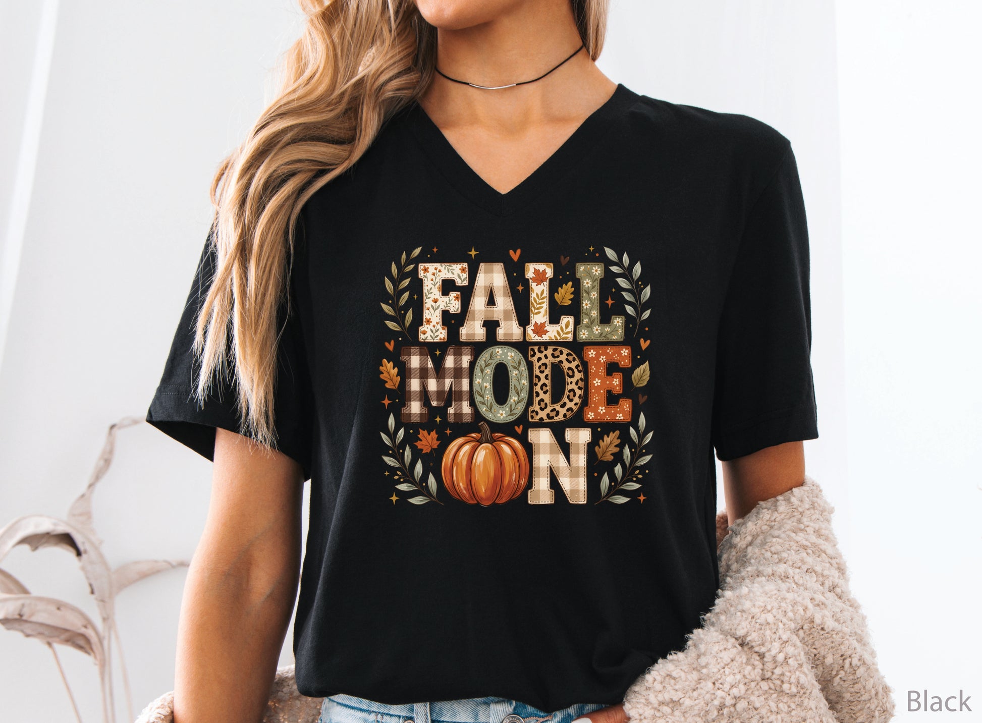 Fall Mode On v-neck t-shirt design featuring patchwork-style lettering with patchwork plaid, floral, leopard print, and fall leaf patterns, plus an orange pumpkin and autumn foliage.