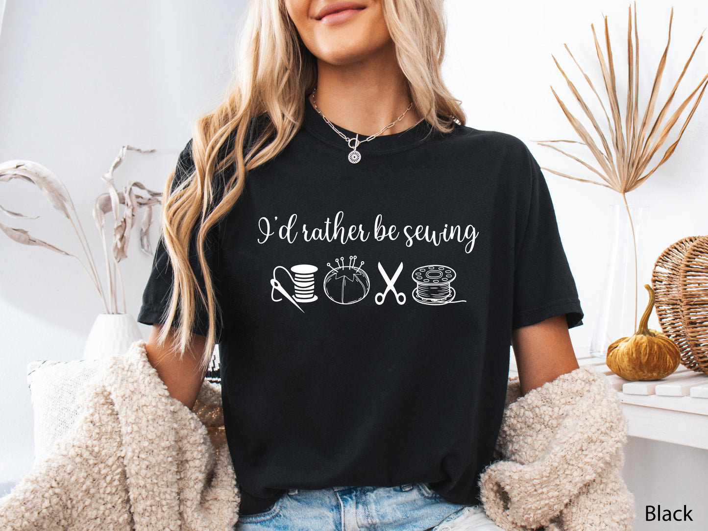 A t-shirt featuring the phrase “I’d Rather Be Sewing” with simple line-art icons of thread spools, a pincushion, scissors, and a needle and thread.