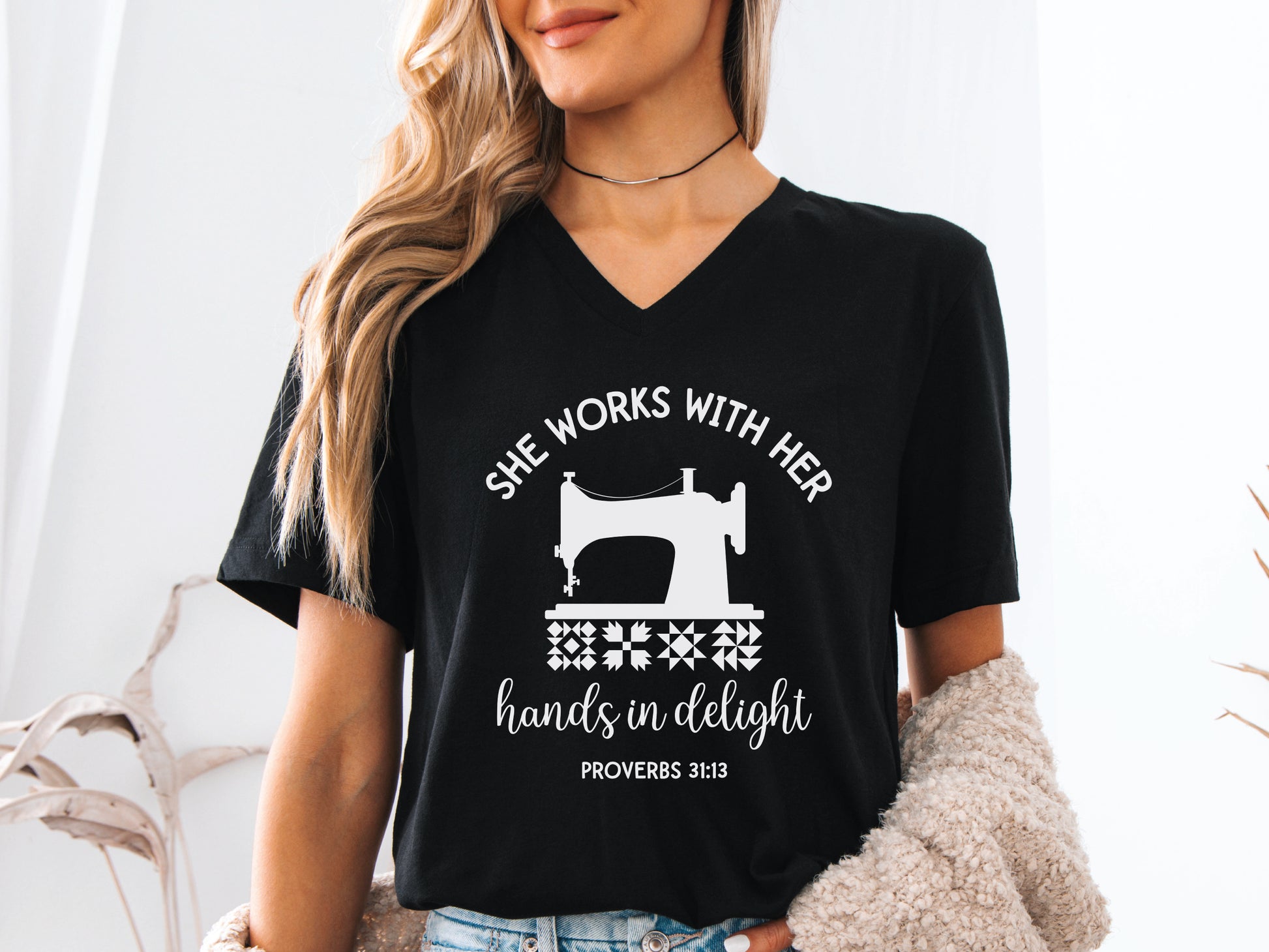 A quilt-themed v-neck  t-shirt featuring a vintage-style sewing machine, quilt block icons, and the verse “She works with her hands in delight – Proverbs 31:13.” 