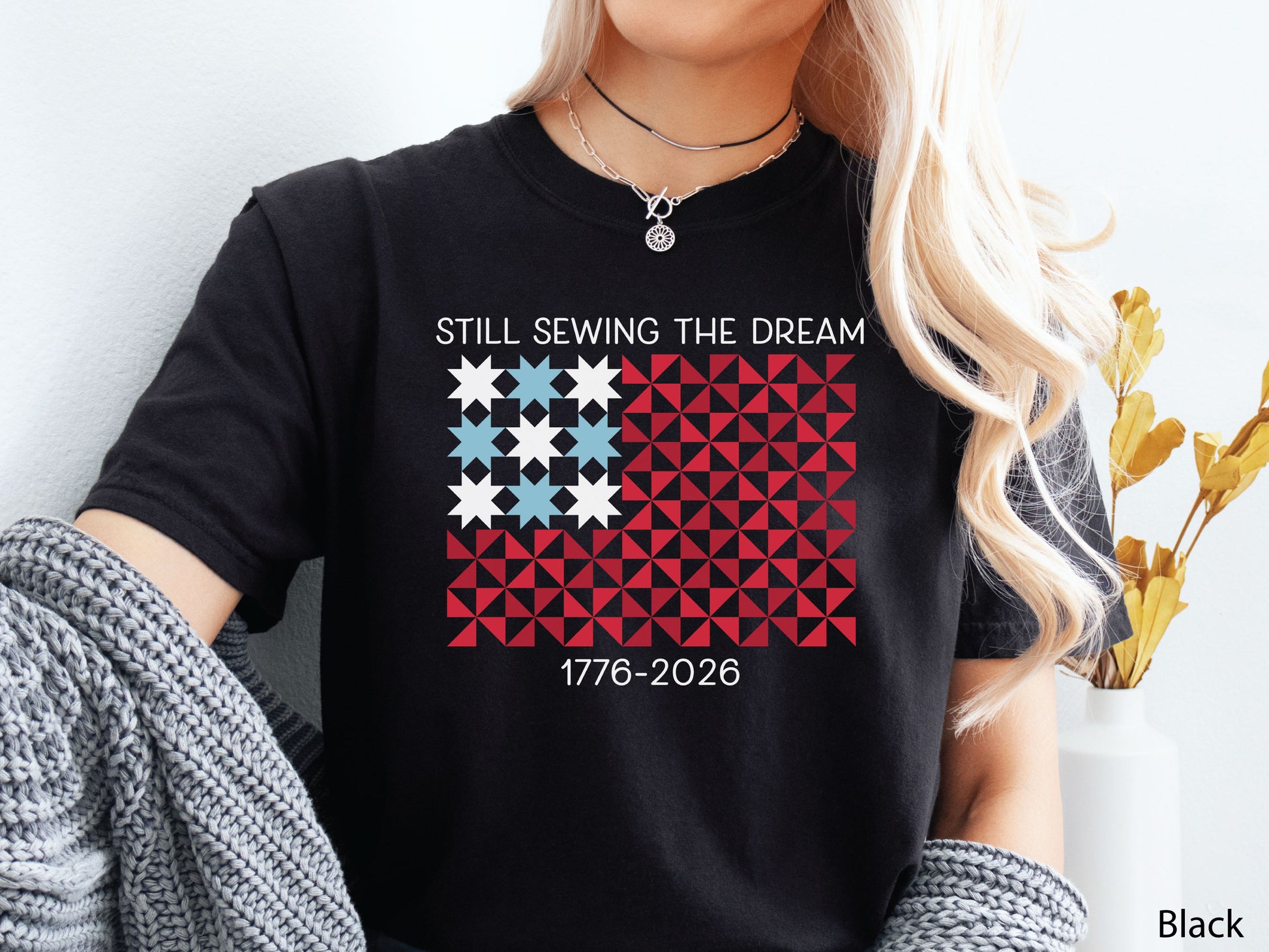 Patriotic quilting v-neck t-shirt with a quilt block American flag made of stars and pinwheel blocks and the phrase “Still Sewing the Dream 1776–2026” in red, white, and blue.
