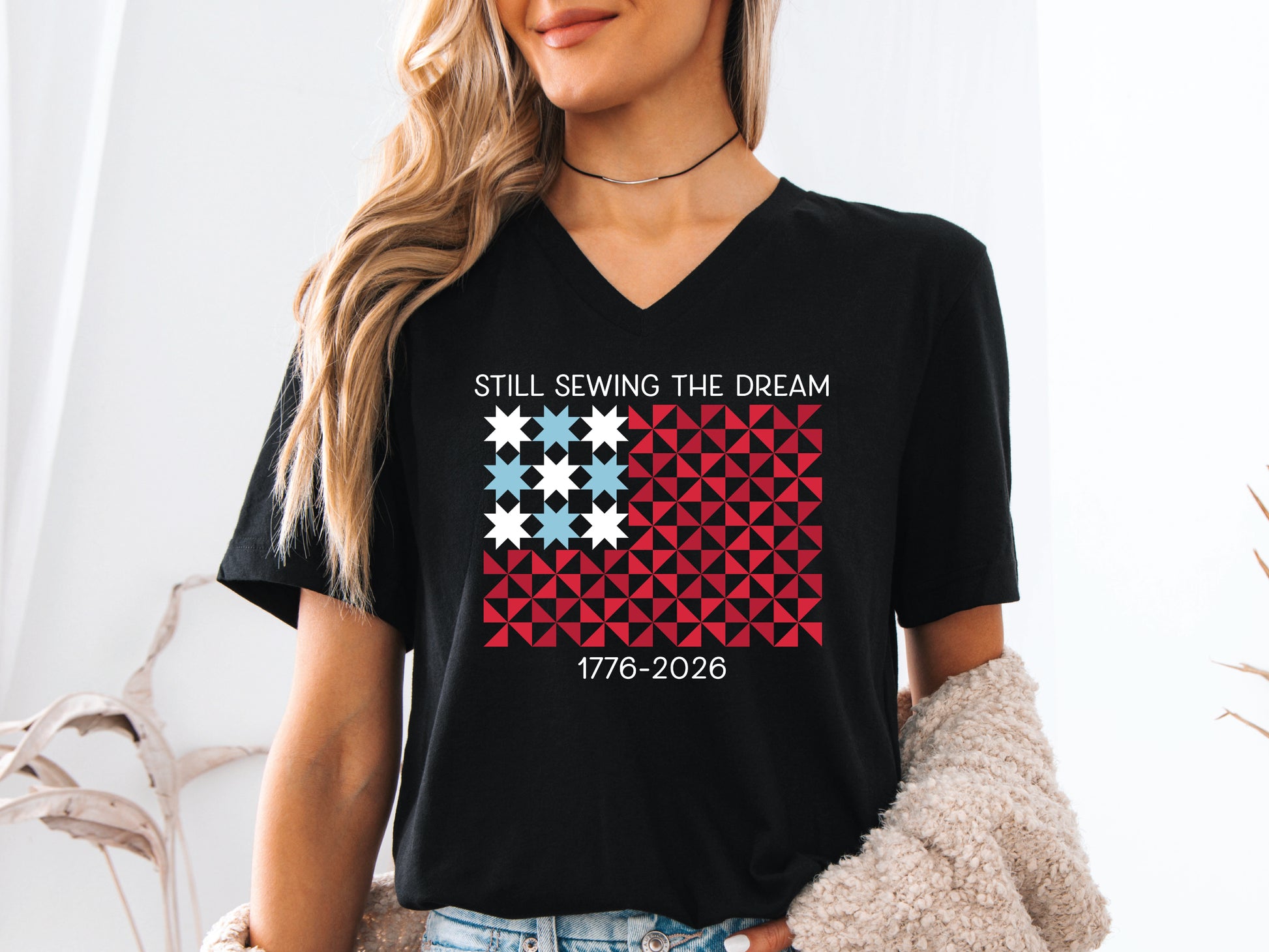 Patriotic quilting v-neck t-shirt with a quilt block American flag made of stars and pinwheel blocks and the phrase “Still Sewing the Dream 1776–2026” in red, white, and blue.