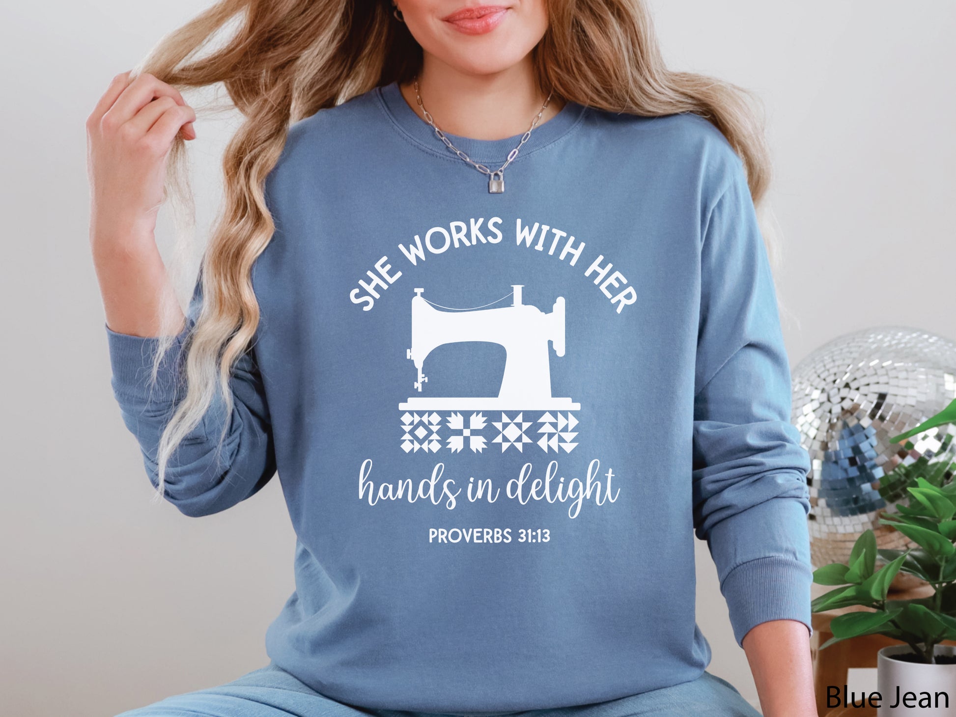 A long-sleeve  t-shirt featuring a vintage-style sewing machine, quilt block icons, and the verse “She works with her hands in delight – Proverbs 31:13.”