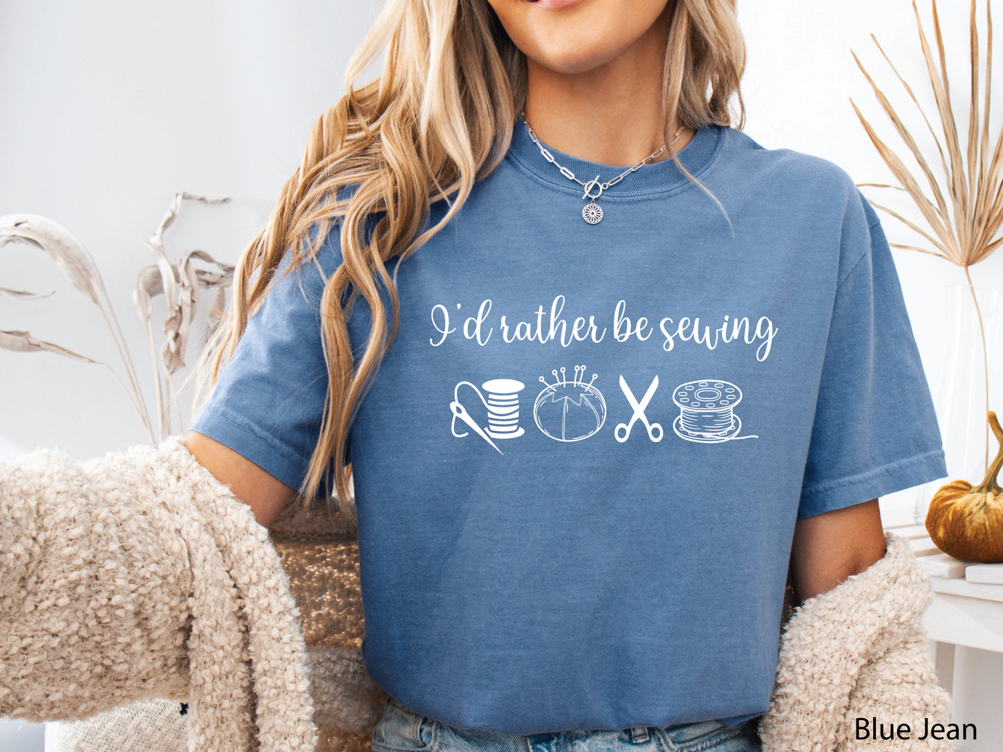 A t-shirt featuring the phrase “I’d Rather Be Sewing” with simple line-art icons of thread spools, a pincushion, scissors, and a needle and thread.