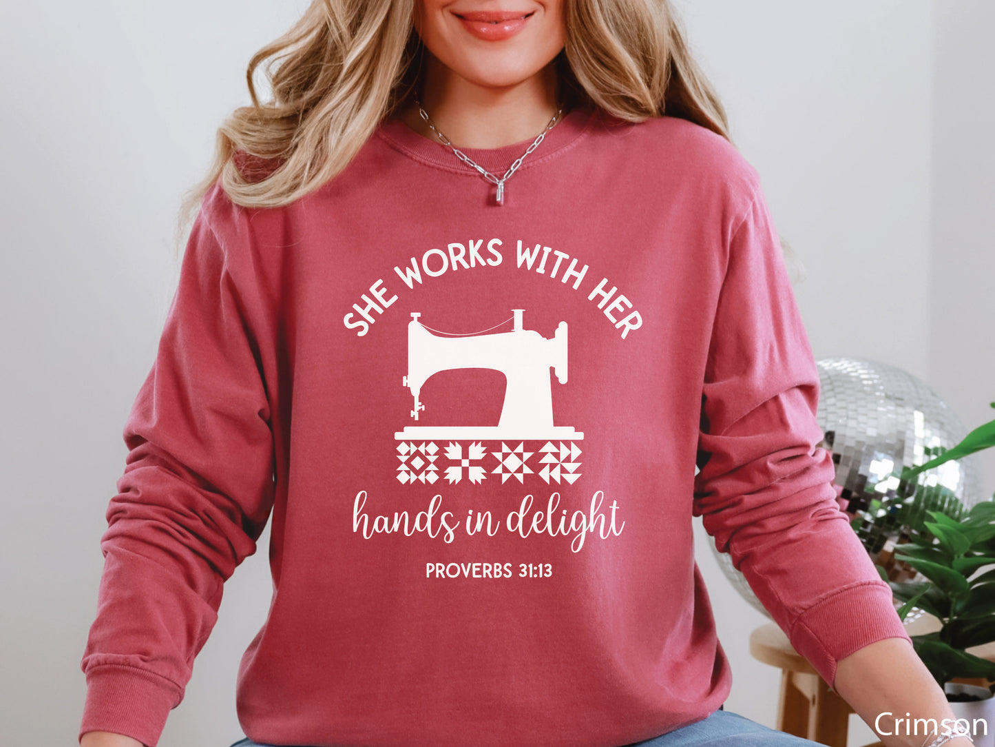 A long-sleeve  t-shirt featuring a vintage-style sewing machine, quilt block icons, and the verse “She works with her hands in delight – Proverbs 31:13.”