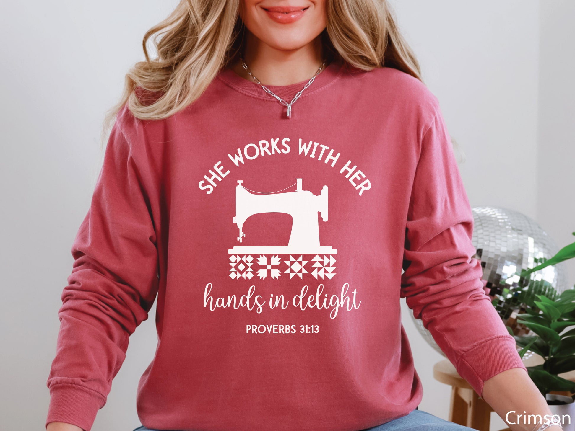 A long-sleeve  t-shirt featuring a vintage-style sewing machine, quilt block icons, and the verse “She works with her hands in delight – Proverbs 31:13.”