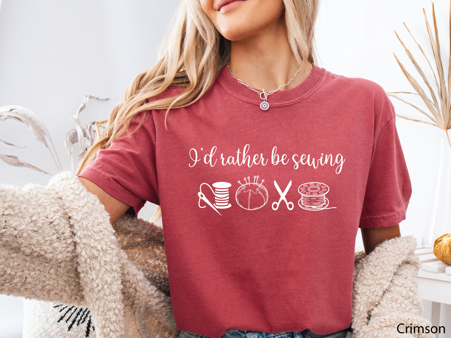 A t-shirt featuring the phrase “I’d Rather Be Sewing” with simple line-art icons of thread spools, a pincushion, scissors, and a needle and thread.