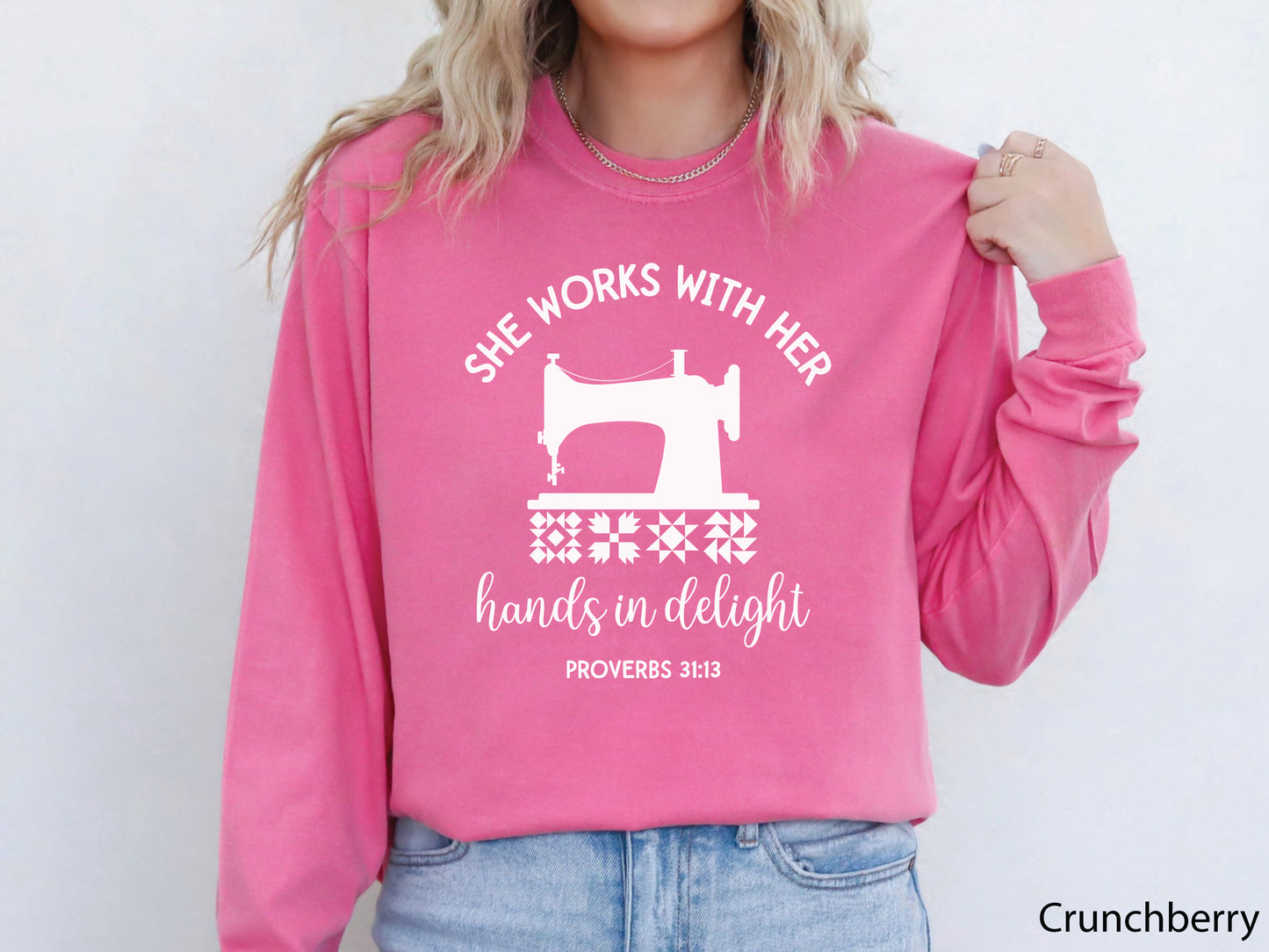 A long-sleeve  t-shirt featuring a vintage-style sewing machine, quilt block icons, and the verse “She works with her hands in delight – Proverbs 31:13.”