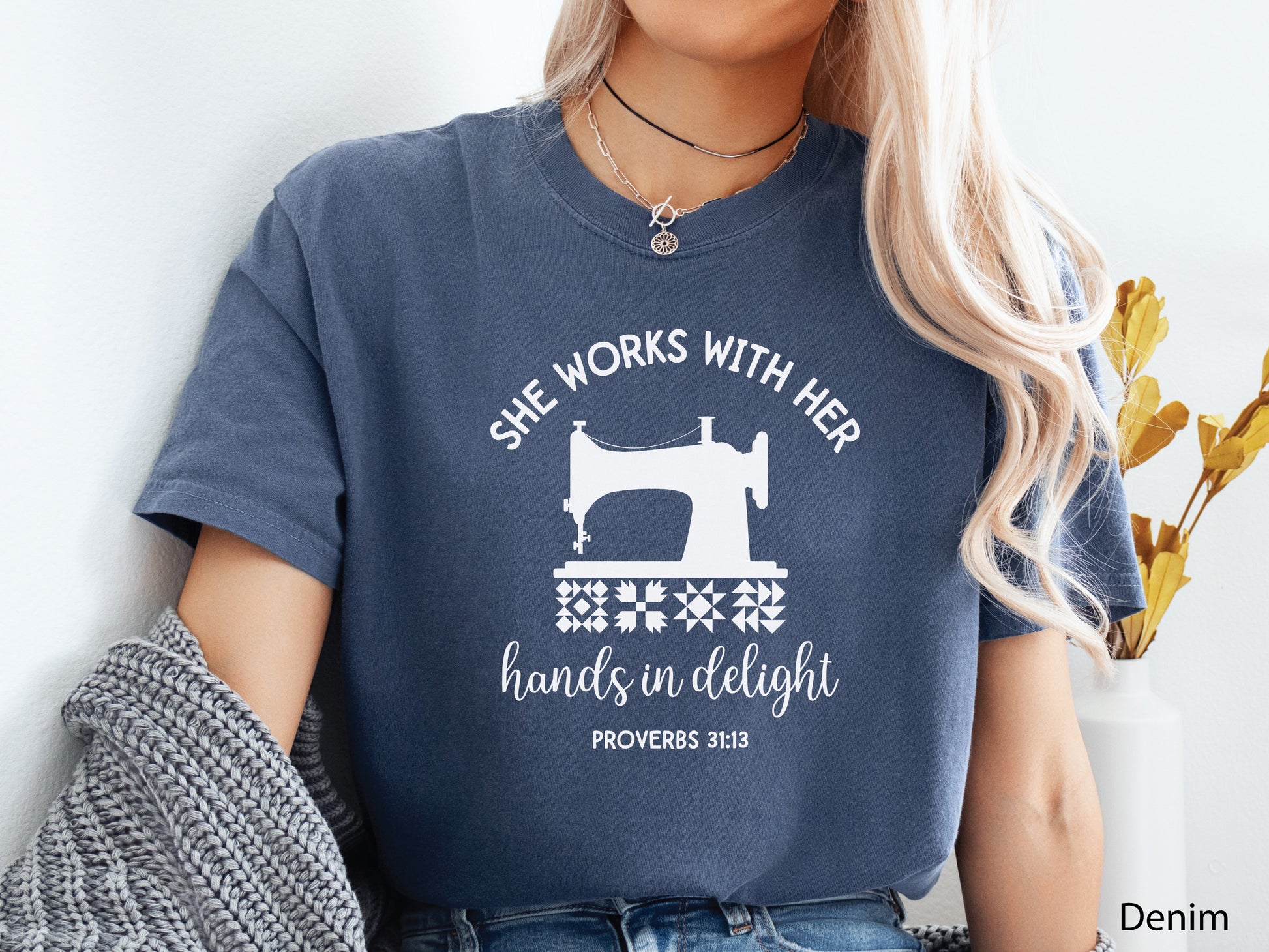 A t-shirt featuring a vintage-style sewing machine, quilt block icons, and the verse “She works with her hands in delight – Proverbs 31:13.”