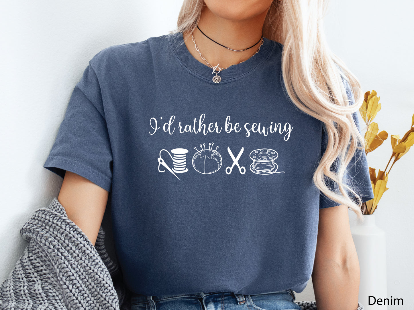 A t-shirt featuring the phrase “I’d Rather Be Sewing” with simple line-art icons of thread spools, a pincushion, scissors, and a needle and thread.