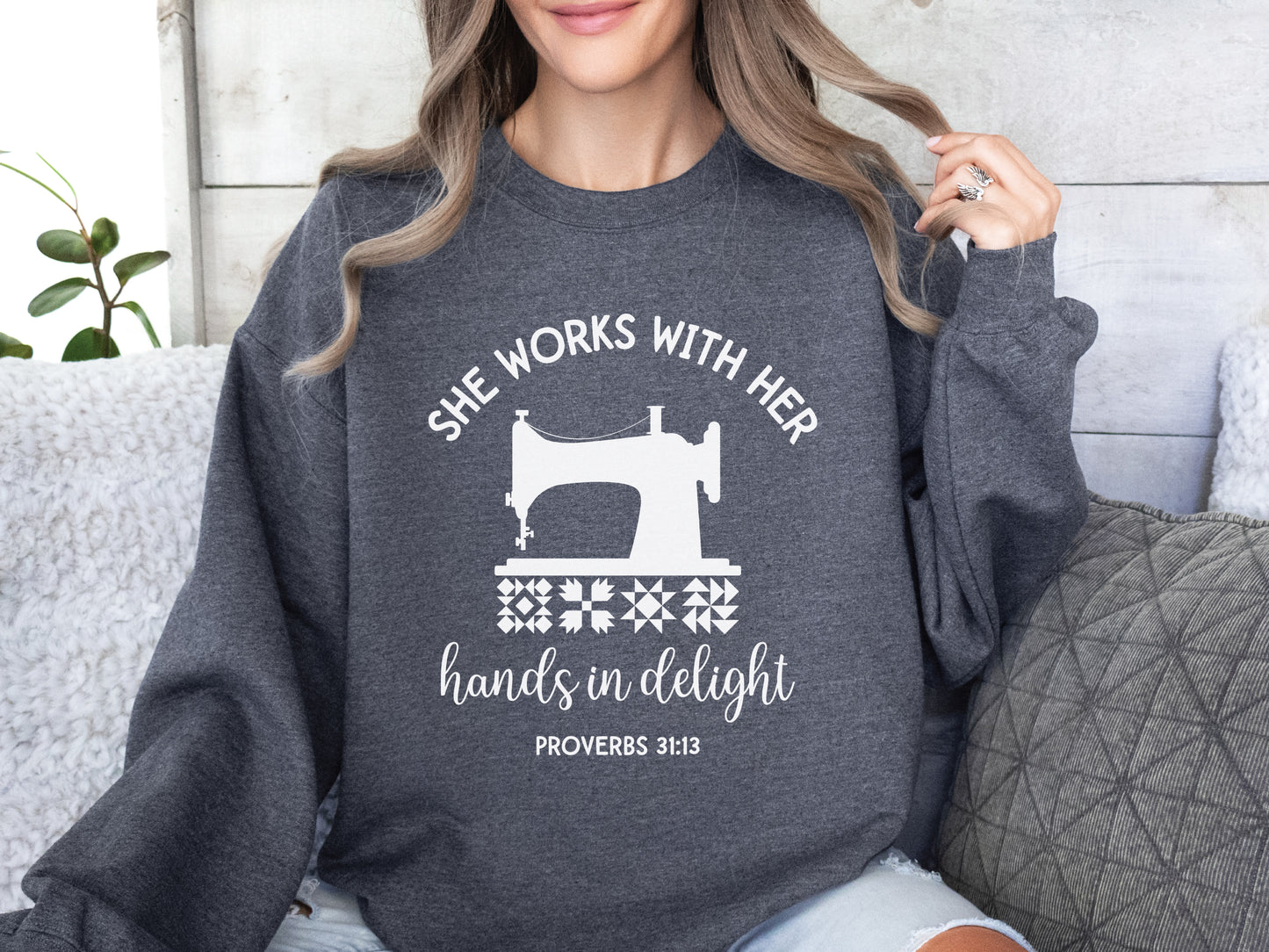 A crew style sweatshirt featuring a vintage-style sewing machine, quilt block icons, and the verse “She works with her hands in delight – Proverbs 31:13.”