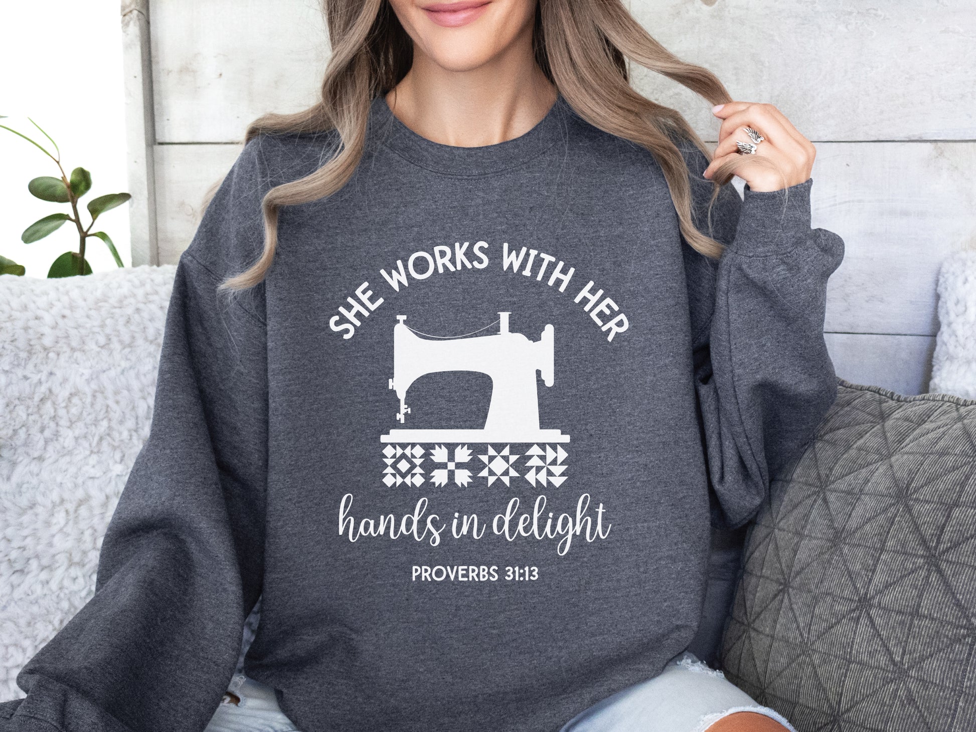 A crew style sweatshirt featuring a vintage-style sewing machine, quilt block icons, and the verse “She works with her hands in delight – Proverbs 31:13.”
