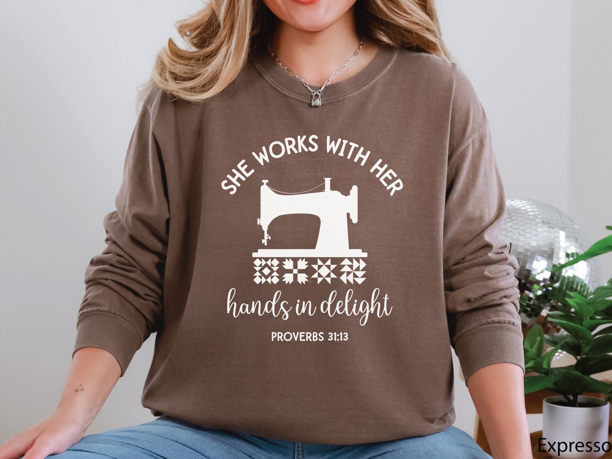 A long-sleeve  t-shirt featuring a vintage-style sewing machine, quilt block icons, and the verse “She works with her hands in delight – Proverbs 31:13.”