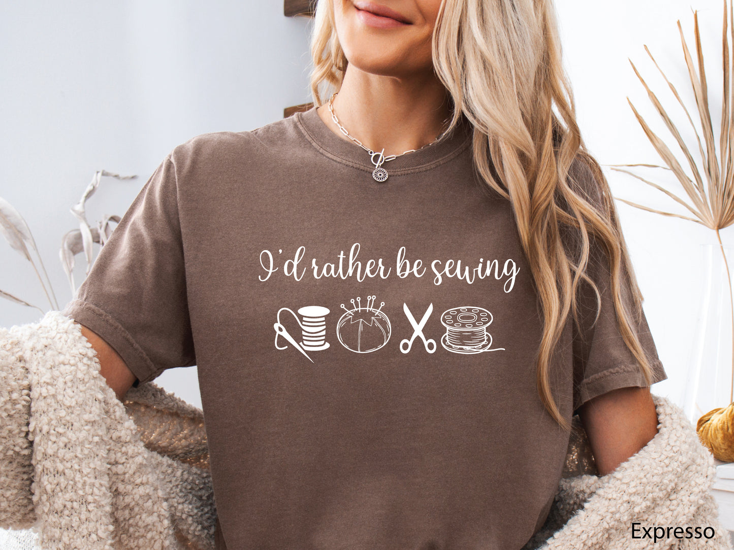 A t-shirt featuring the phrase “I’d Rather Be Sewing” with simple line-art icons of thread spools, a pincushion, scissors, and a needle and thread.